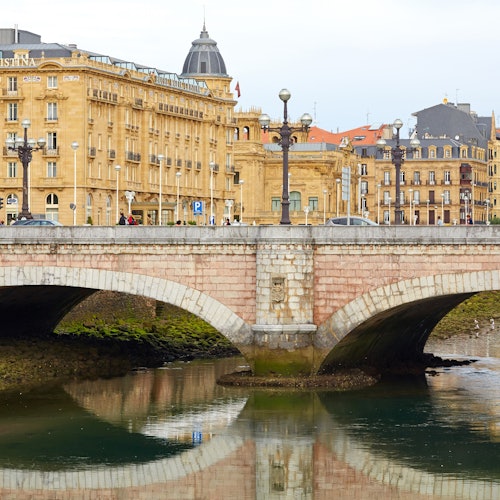 People walking across a stone bridge with arched openings over a calm river, with historic yellow buildings in the background.