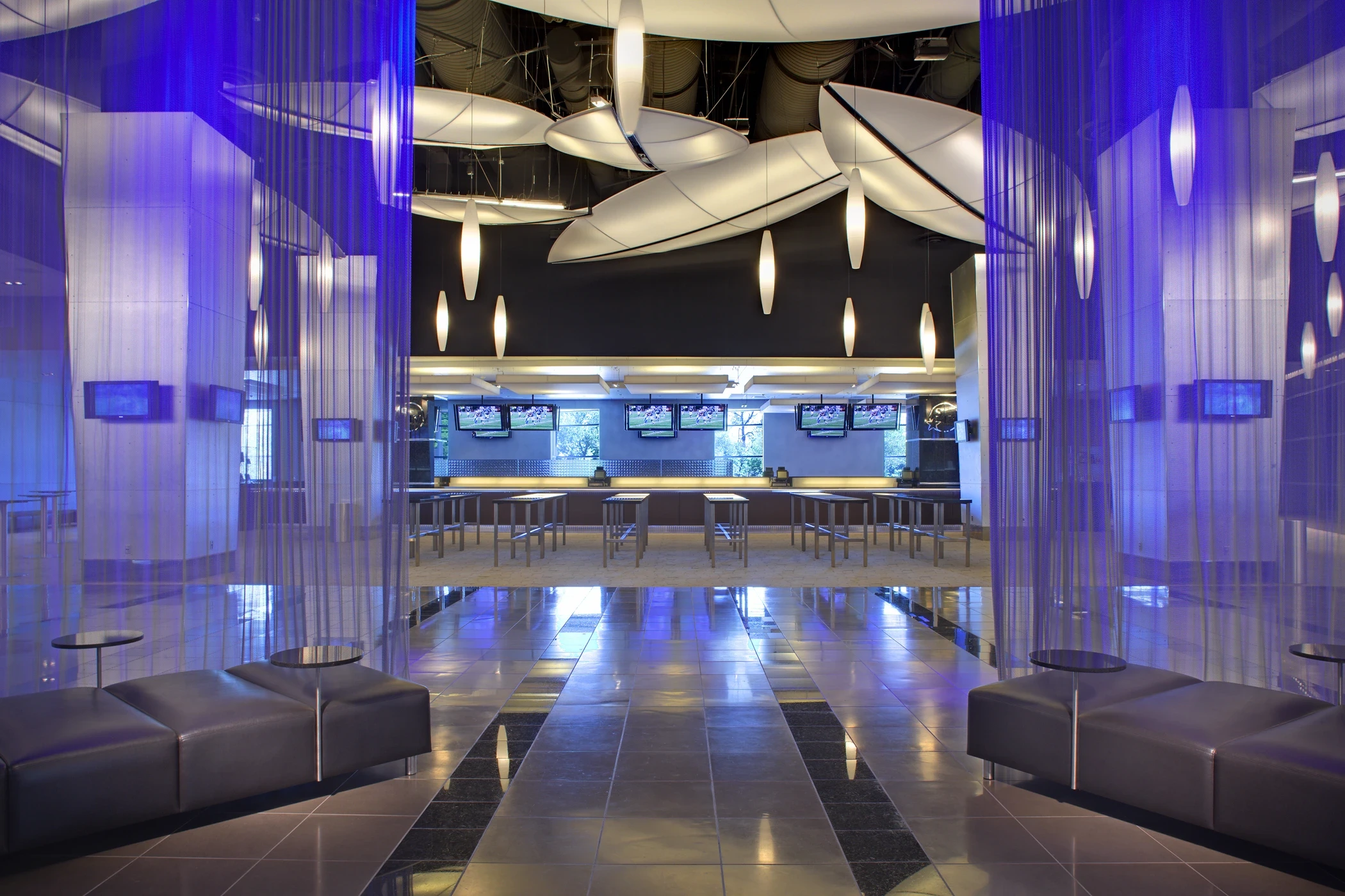 Modern stadium club lounge with purple lighting, sleek seating, high ceilings, and multiple televisions above a bar area.