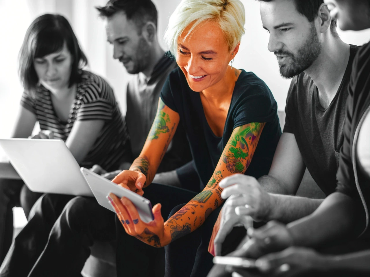 Group of people collaborating with tablets and laptops, featuring a tattooed woman at the center.