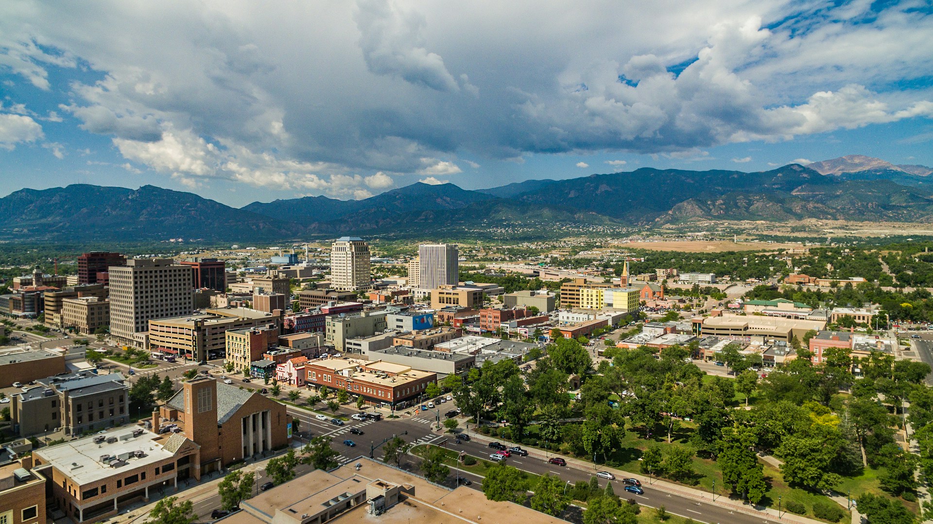 Downtown skyline with mid-rise office buildings backed by forested mountains under cloudy skies.
