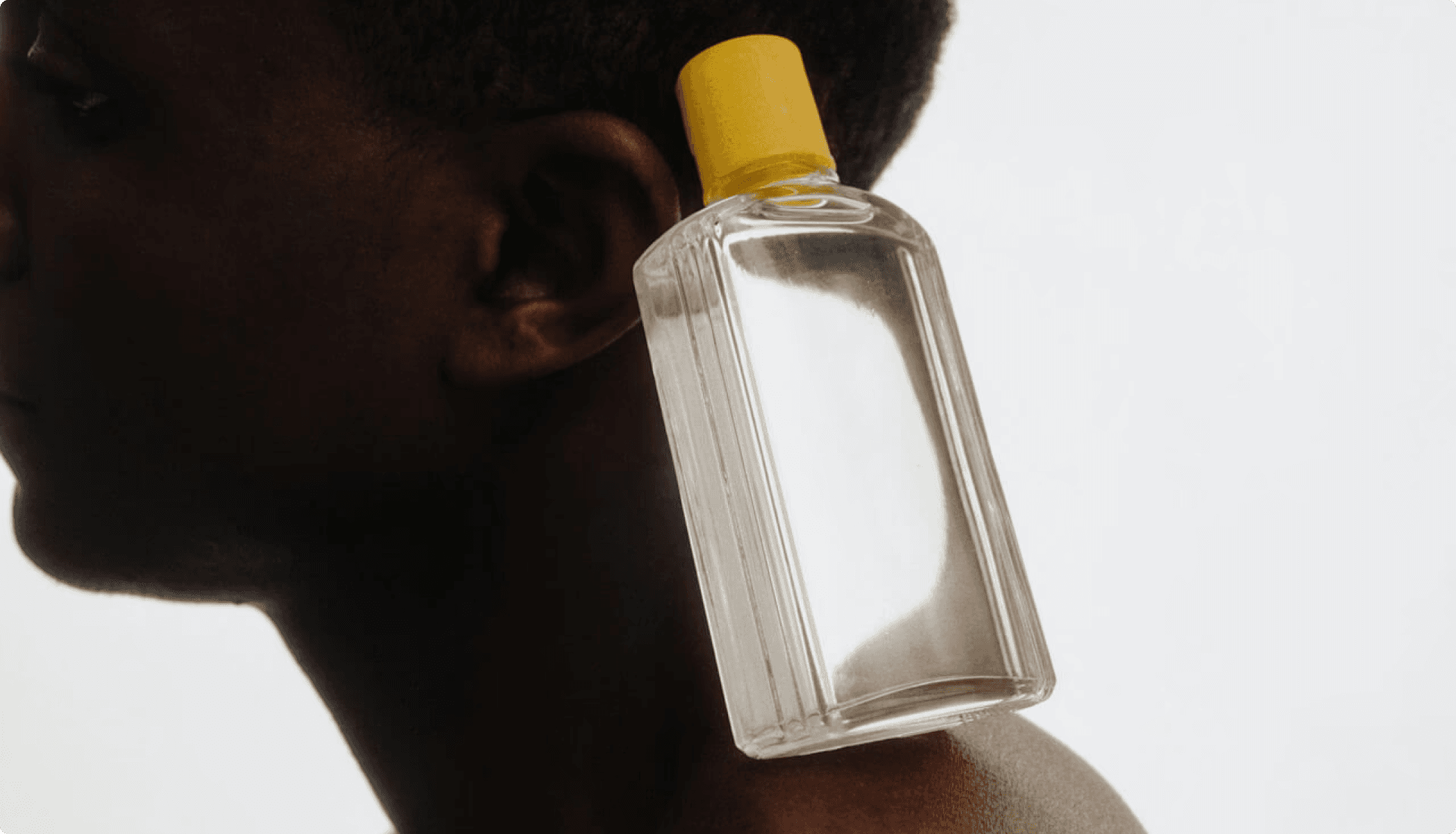 A minimalist composition featuring a clear glass bottle with a yellow cap balanced on a person’s shoulder against a white background.