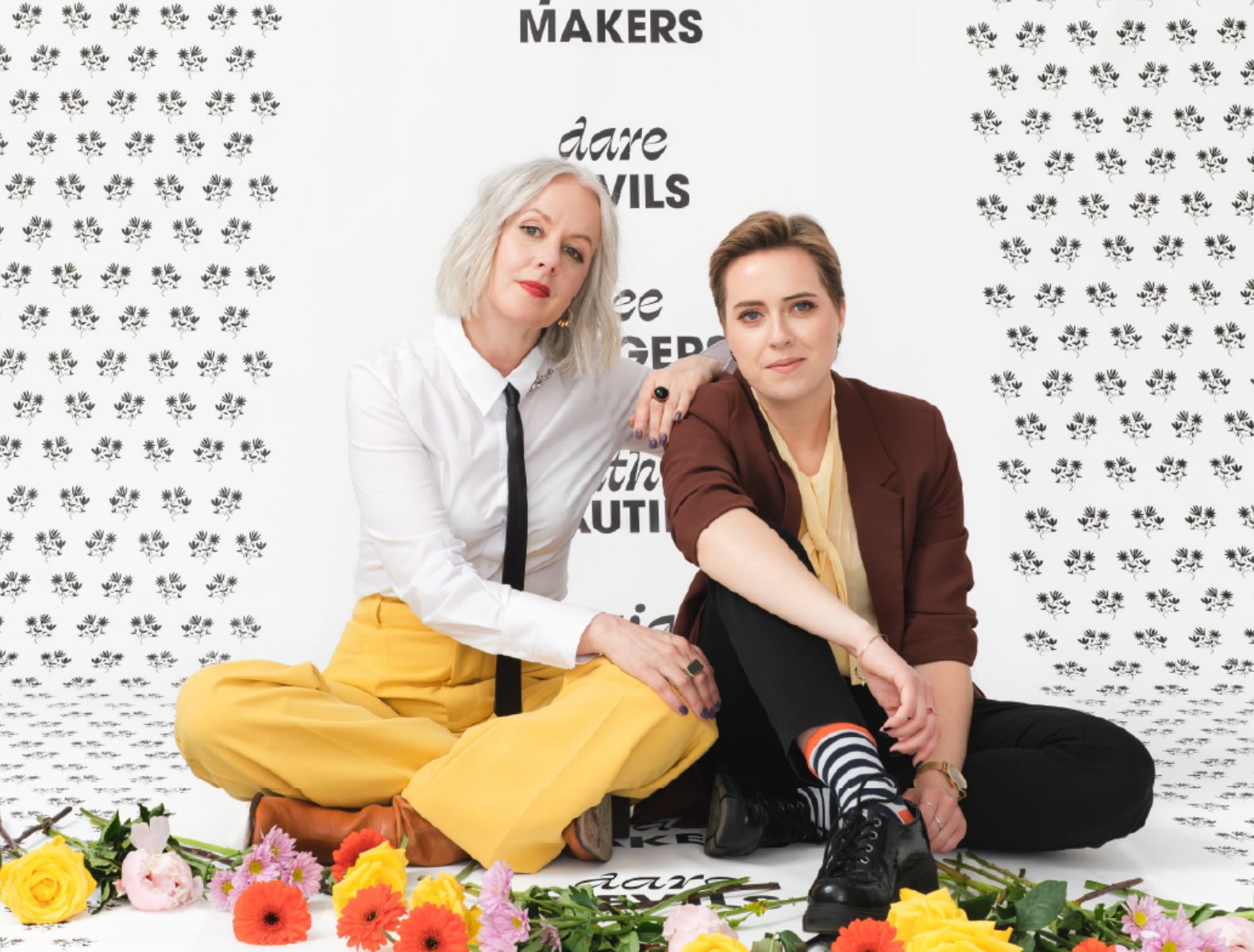 Emily Kiana and Liz parsons in dress pants and button up shirts sitting on a branded background that says "dare devils" surrounded by yellow, pink, and orange cut flowers. 