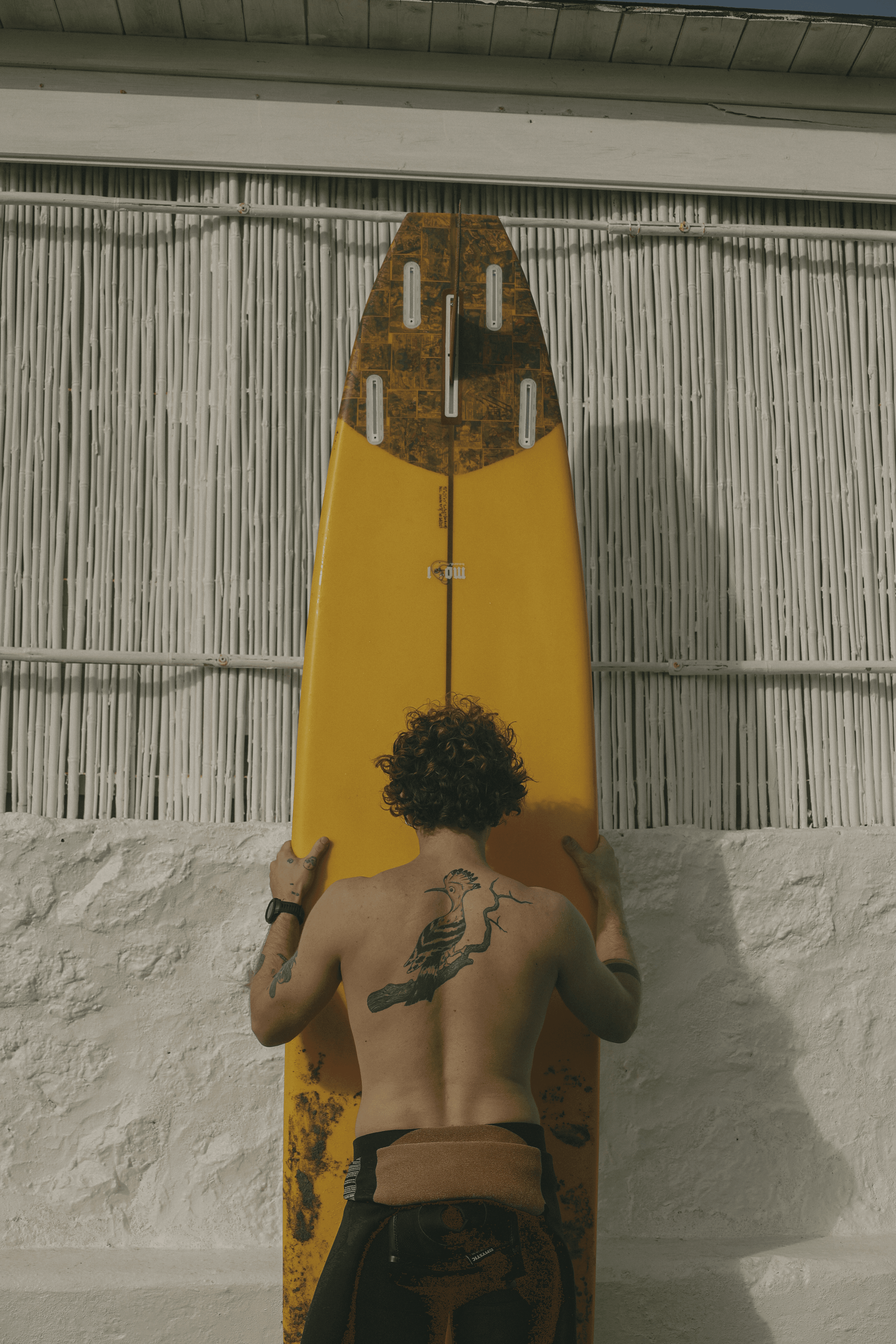 Vista posteriore di un uomo a torso nudo e tatuato che tiene in verticale una longboard da surf giallo brillante con un pattern verde-marrone sulla coda.