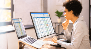A woman sits in front of a laptop and a large desktop monitor. Both screens show spreadsheets.
