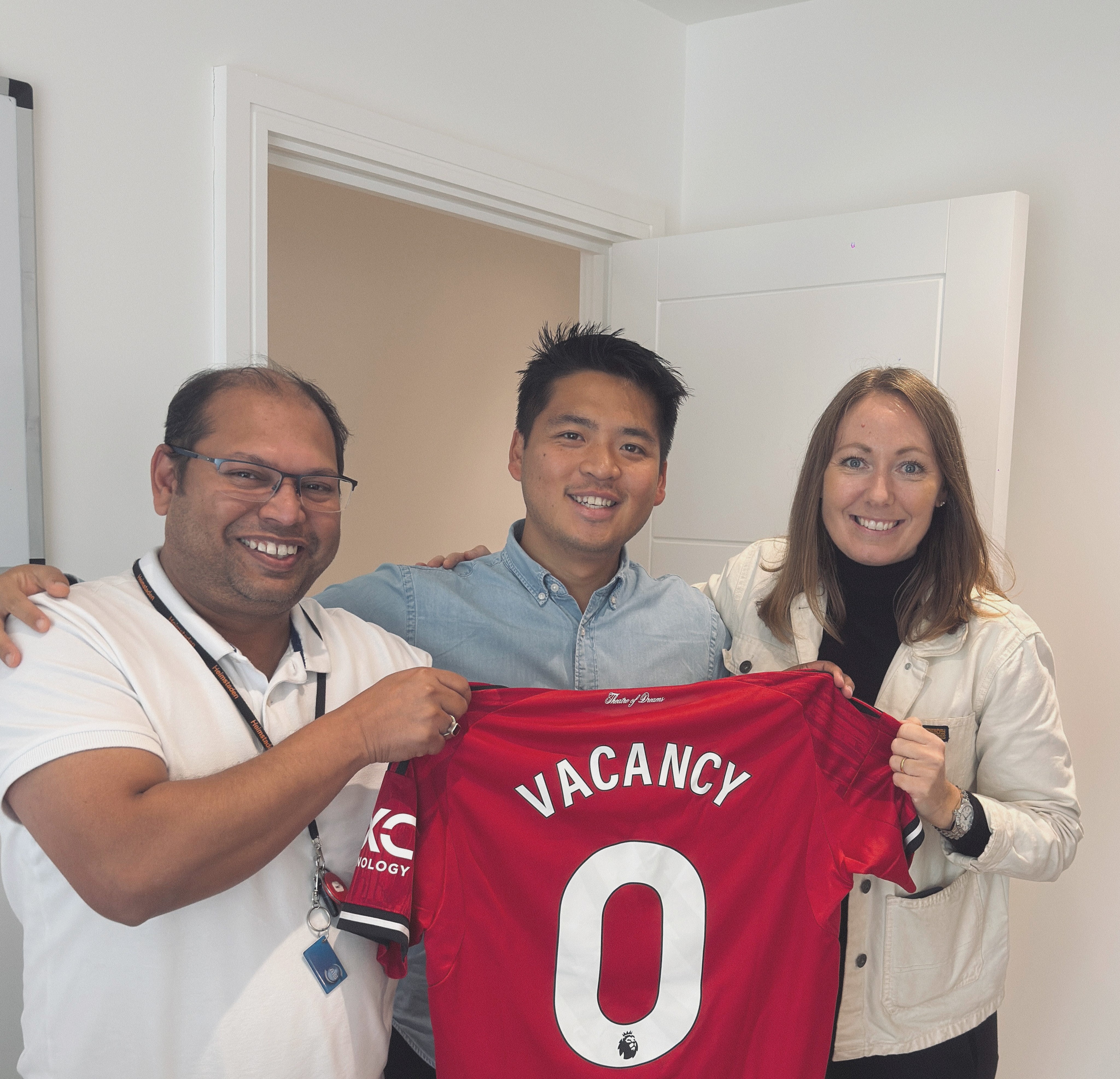 Three smiling people holding a red Manchester United jersey printed with 'VACANCY 0