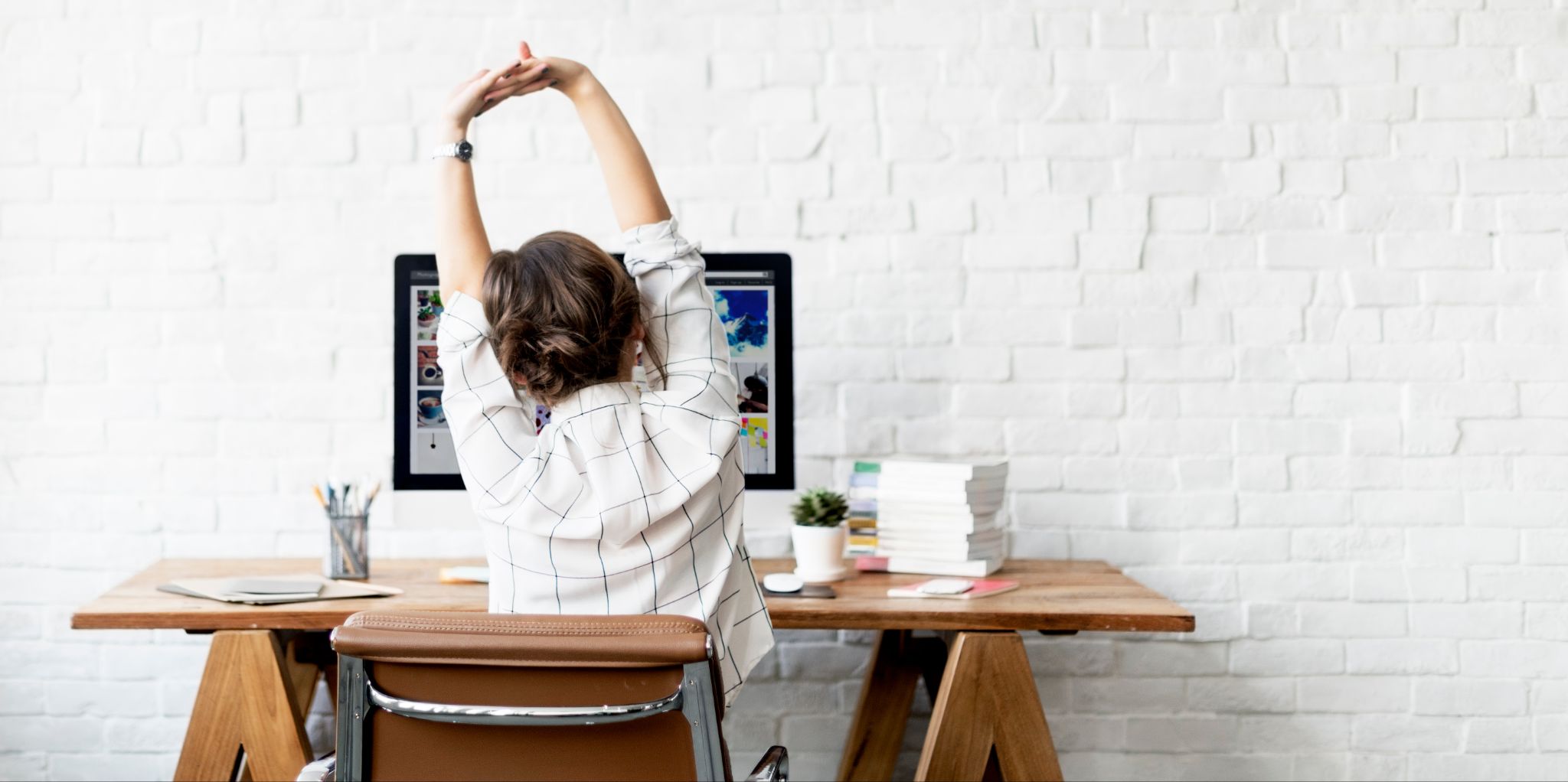 A woman sitting at a wooden desk stretching her arms overhead while working on a computer in a bright office with a white brick wall.