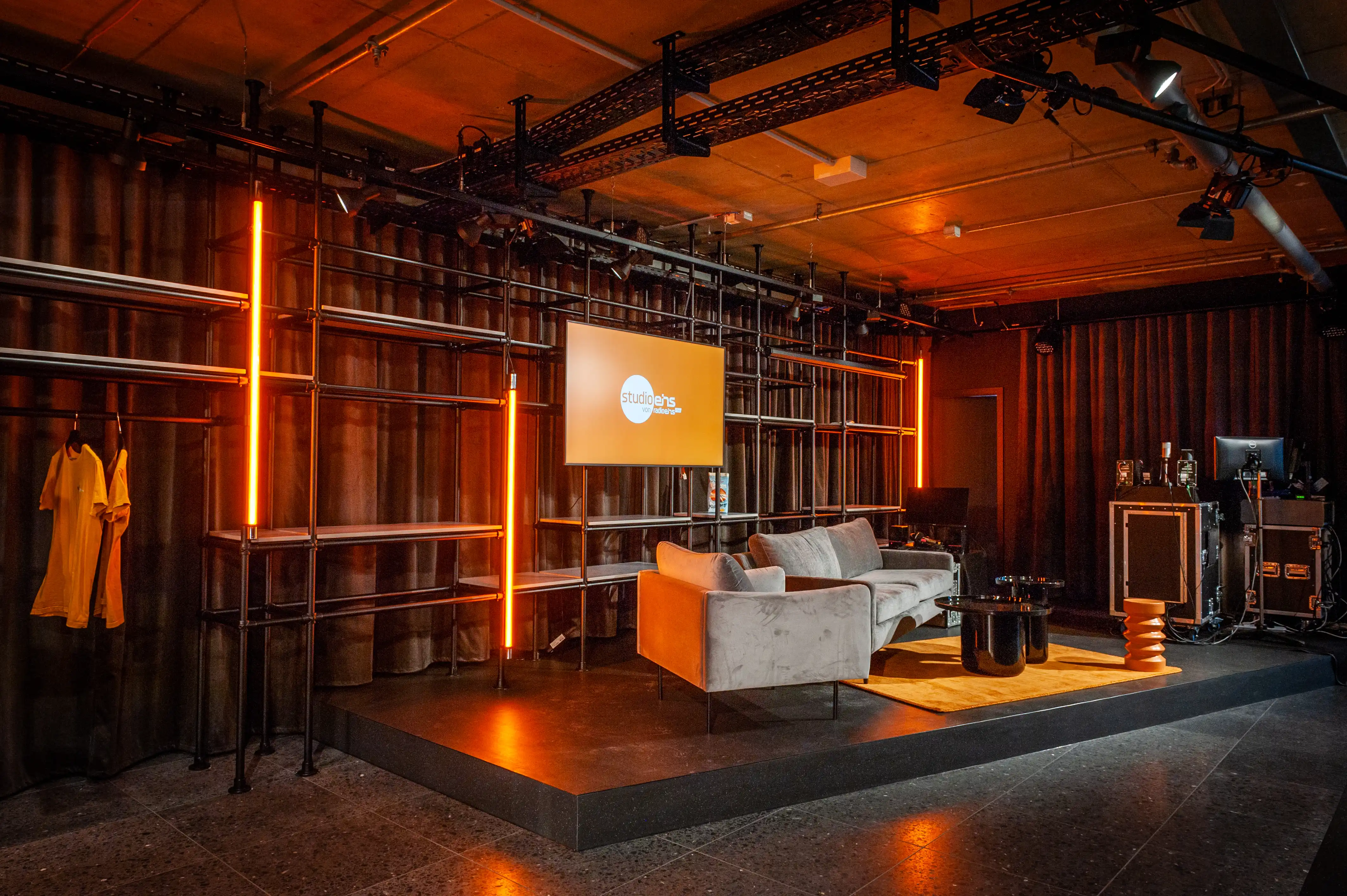 Stage set of the Radio Eins studio with grey sofa, screen and orange light lines in front of black scaffold shelving.