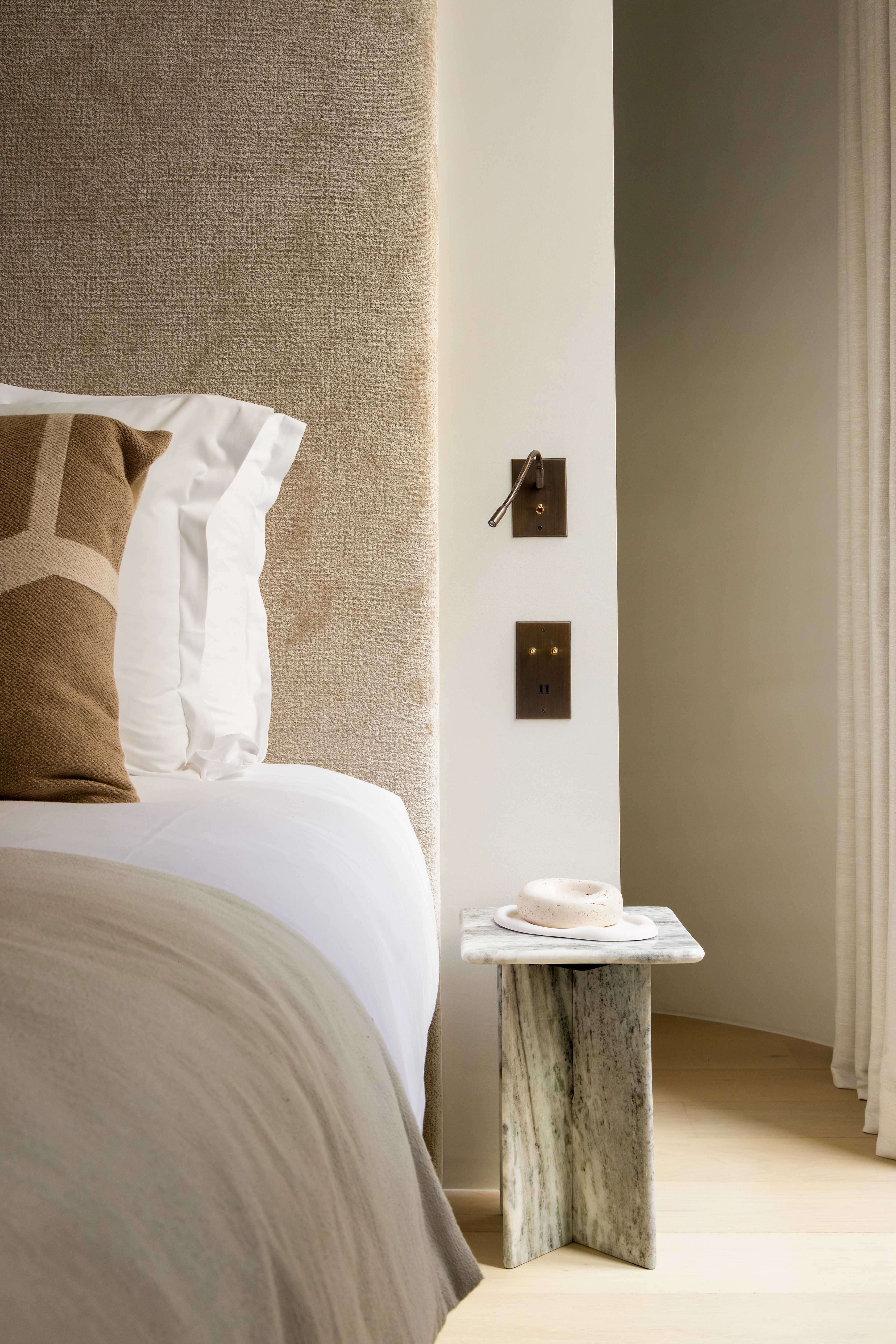 Detail de la tete de lit en velours beige avec coussins graphiques, liseuse doree et table de chevet en bois brut
