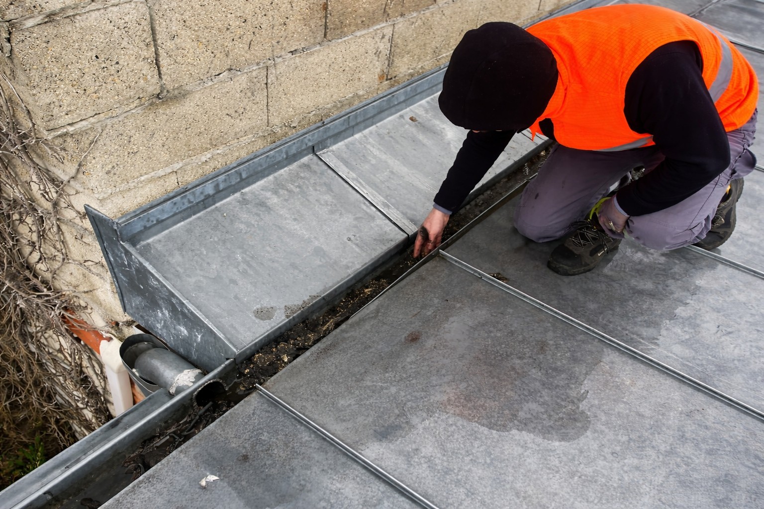 Technicien en gilet orange nettoyant un chéneau en zinc sur toiture plate, avec débris visibles près de la descente d’eaux pluviales