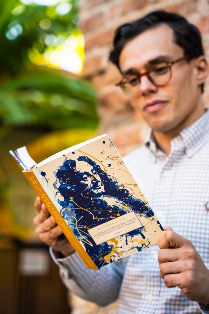 A woman with long hair and a black blazer holds a copy of Boethius's "The Consolation of Philosophy" next to her face, published by Logos.