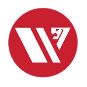 Smiling young woman with curly hair sitting at a laptop, in front of a teal wall.