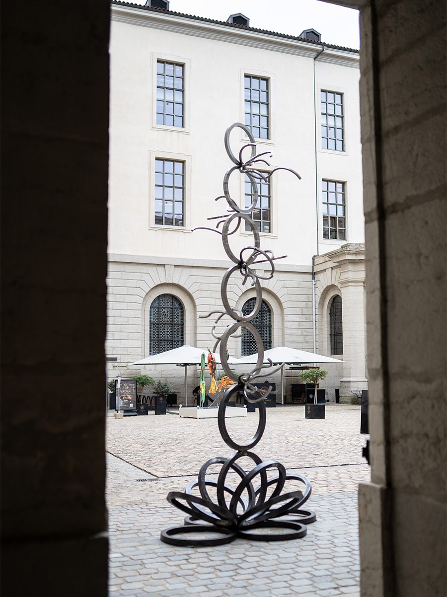Une sculpture métallique verticale composée d'anneaux empilés est installée au centre d'une cour pavée, vue à travers une ouverture encadrée de pierre. La base de l'œuvre forme une structure circulaire complexe, tandis que la partie supérieure s'élève avec des cercles entrelacés et des éléments fins et incurvés. En arrière-plan, la cour est délimitée par un bâtiment classique à plusieurs étages avec des fenêtres cintrées et des parasols blancs installés à l'extérieur.