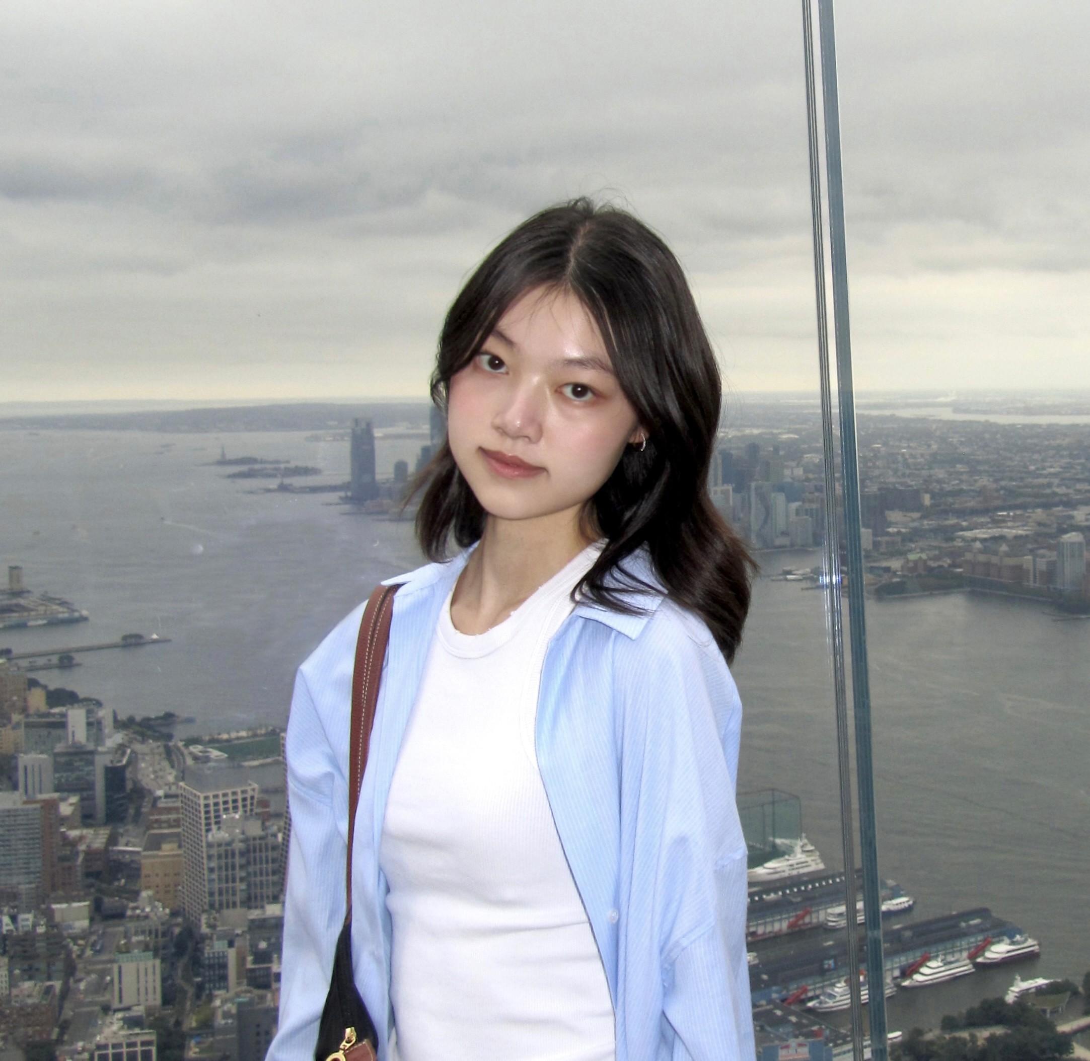 A person stands outdoors in a natural setting with lush green trees under a clear sky, wearing a dark sweatshirt, and looking into the distance.