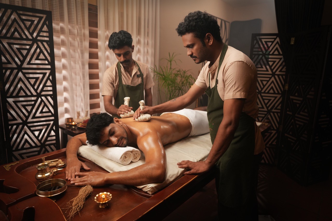 Ayurvedic body purification and detox therapy – A man receiving a synchronized Pinda Sweda (herbal pouch massage) from two therapists in a traditional wellness spa setting.
