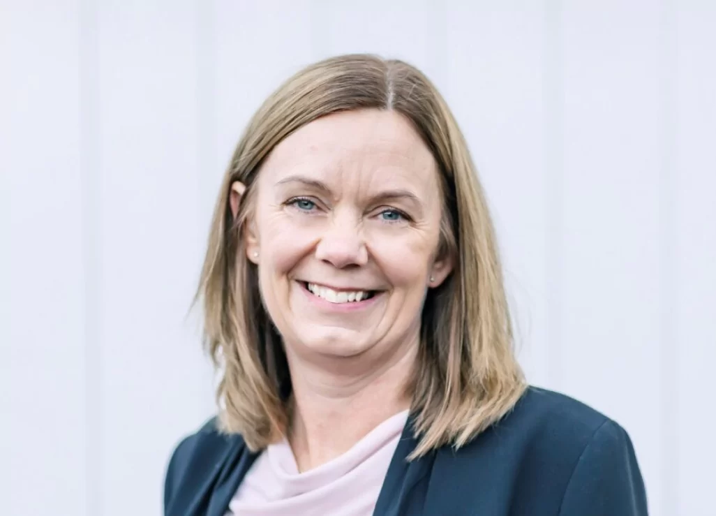 En kvinna med blont hår och ett leende iförd en mörkblå kavaj och en rosa blus.