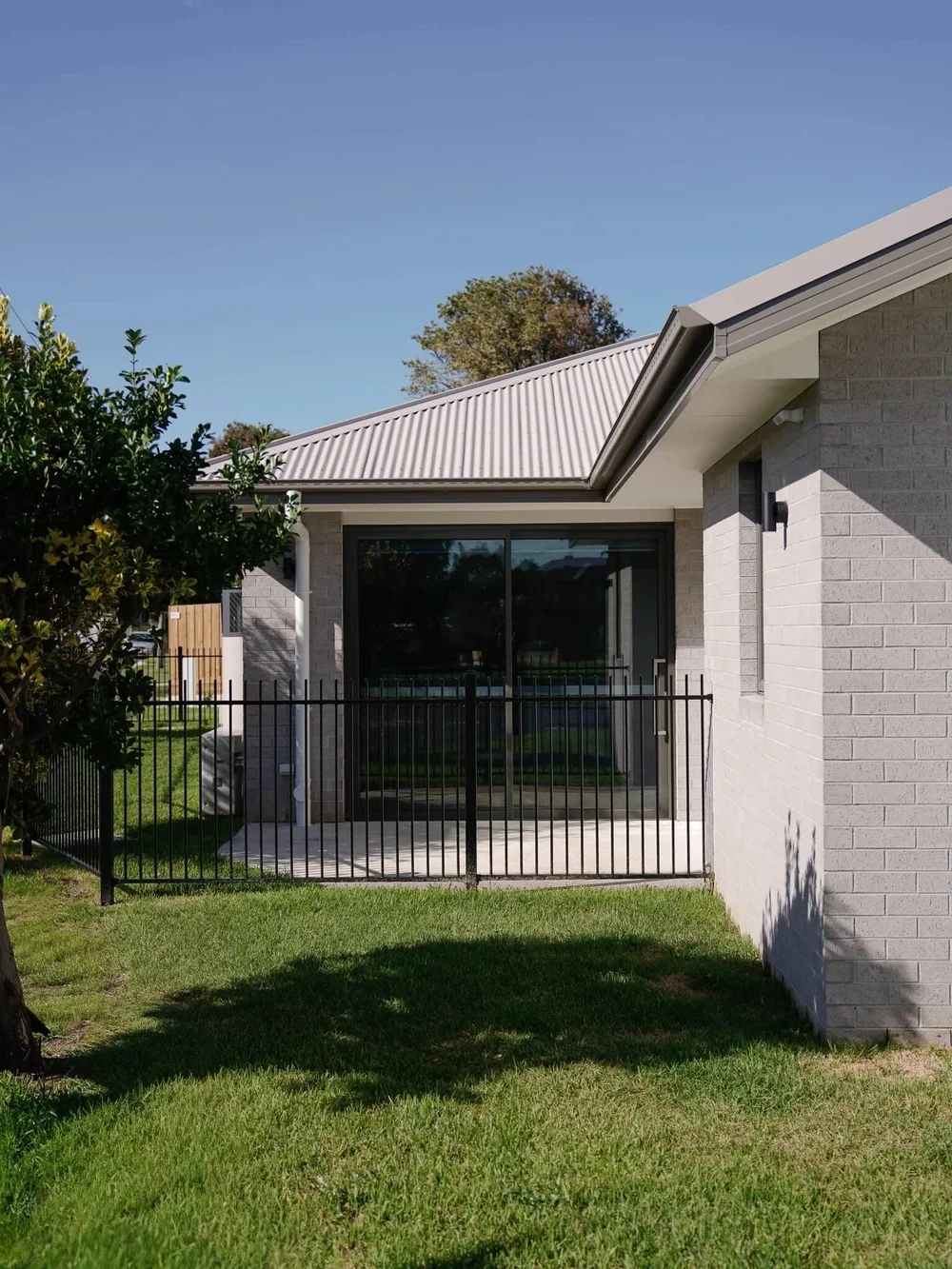 Modern gray brick house with black fence and green lawn under clear blue sky.