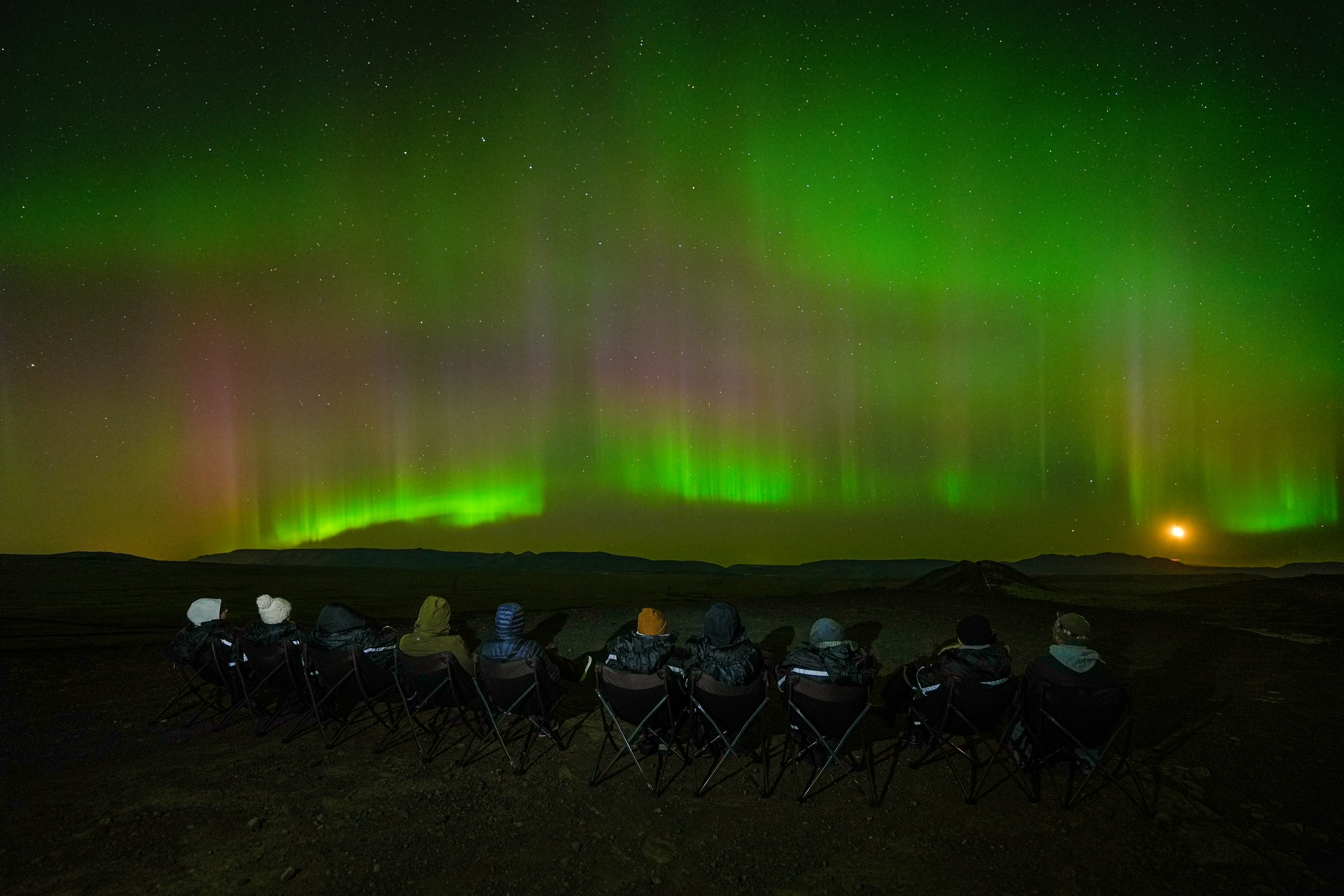 A row of guests in camping chairs watching a wide green and pink aurora display