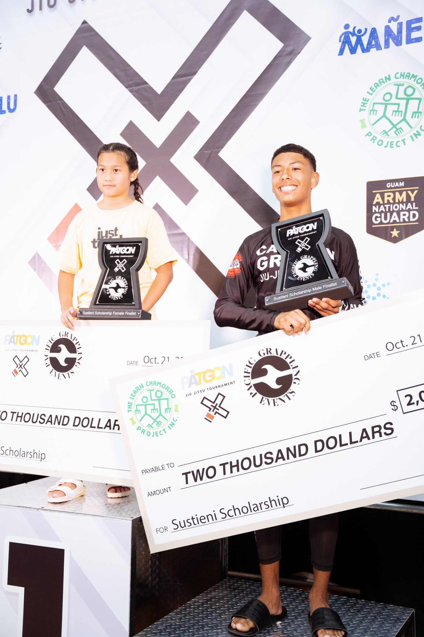 Two young jiu-jitsu competitors hold large checks and trophies on the winner's podium at a Pacific Grappling Events tournament, with branding and sponsor logos in the background.