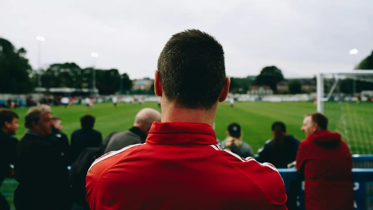 A successful coach in a red training suit attentively watches a football match on a green pitch, surrounded by spectators. The image illustrates the dedication and focus that are crucial for success in high-performance sport.