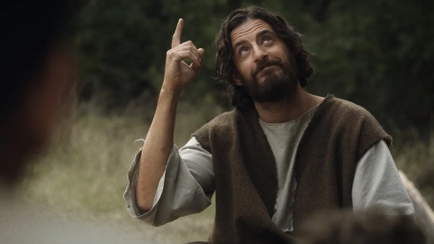A speaker on stage, illuminated against a dark background, gestures while addressing an audience.