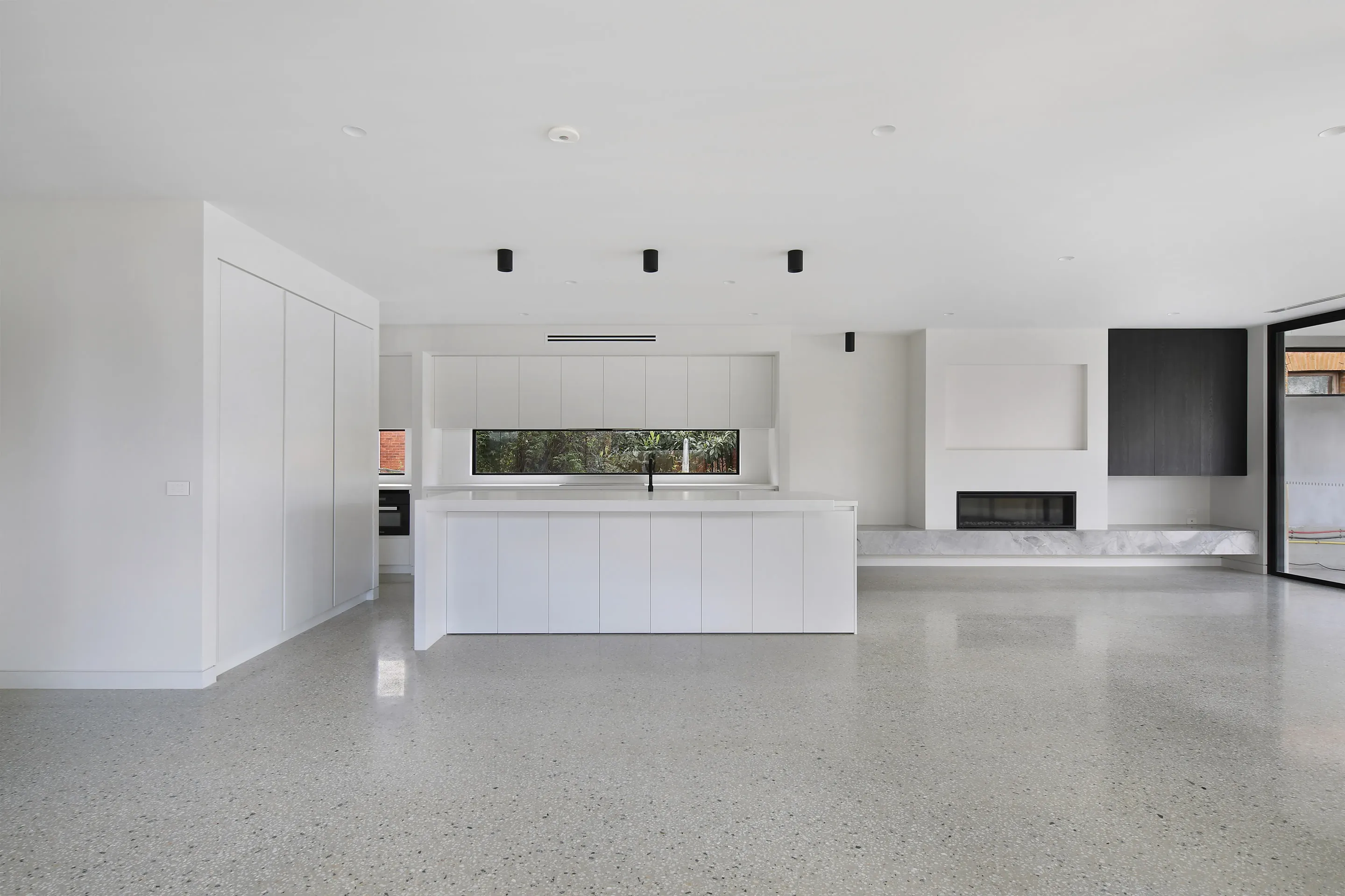 Open-plan kitchen and living with a white waterfall island and black tap, white cabinetry under a long window, a recessed gas fireplace with grey marble bench, black cabinetry and polished-concrete floors
