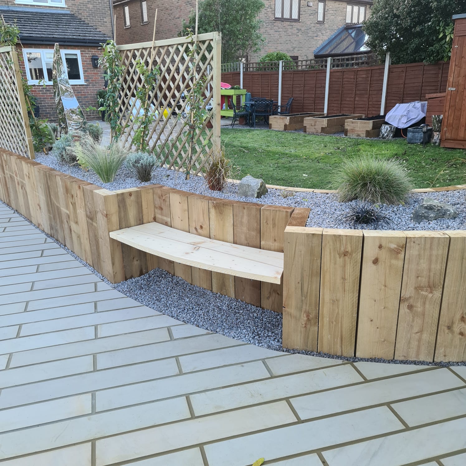 A wooden bench surrounded by low wooden walls and plants in a garden setting. Paved surface visible in foreground.