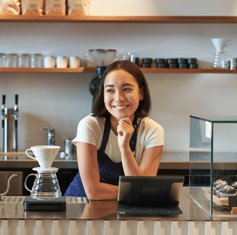 A young person with curly hair smiles at the camera while sitting in front of a laptop against a dark background.