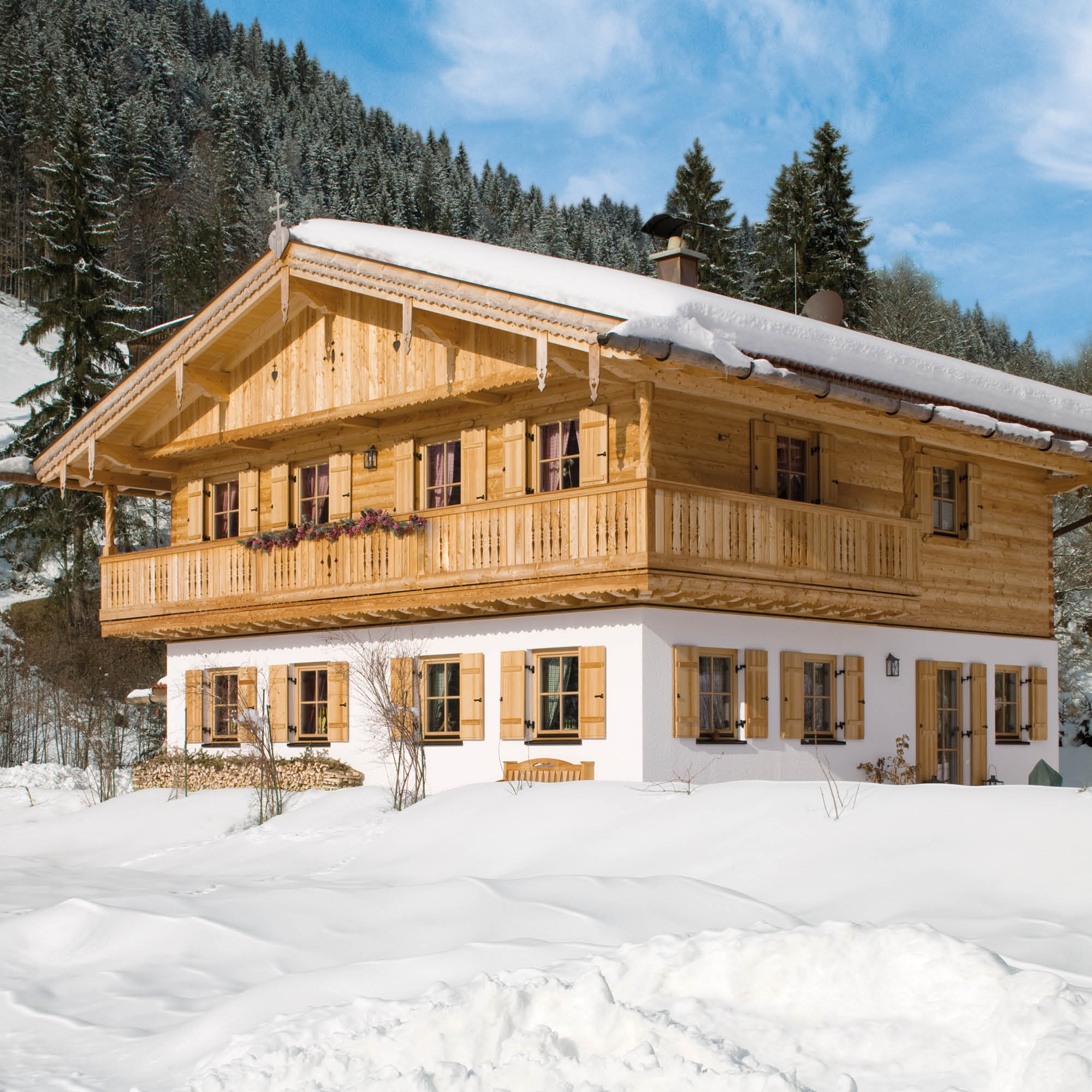 Holzhaus im traditionellen bayerischen Stil mit weißen Fensterläden und Balkon, umgeben von Schnee und Nadelwald.