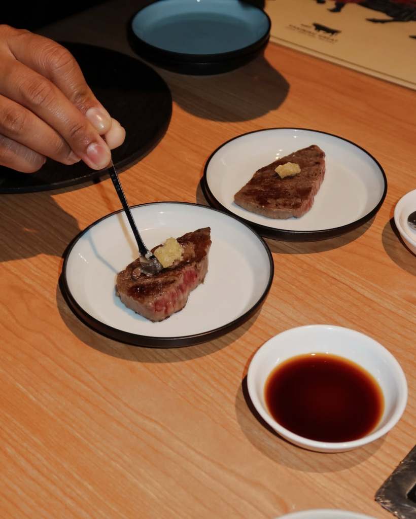 A close-up of a person using a fork to add a topping to a slice of grilled beef on a small plate, accompanied by a small bowl of dipping sauce.
