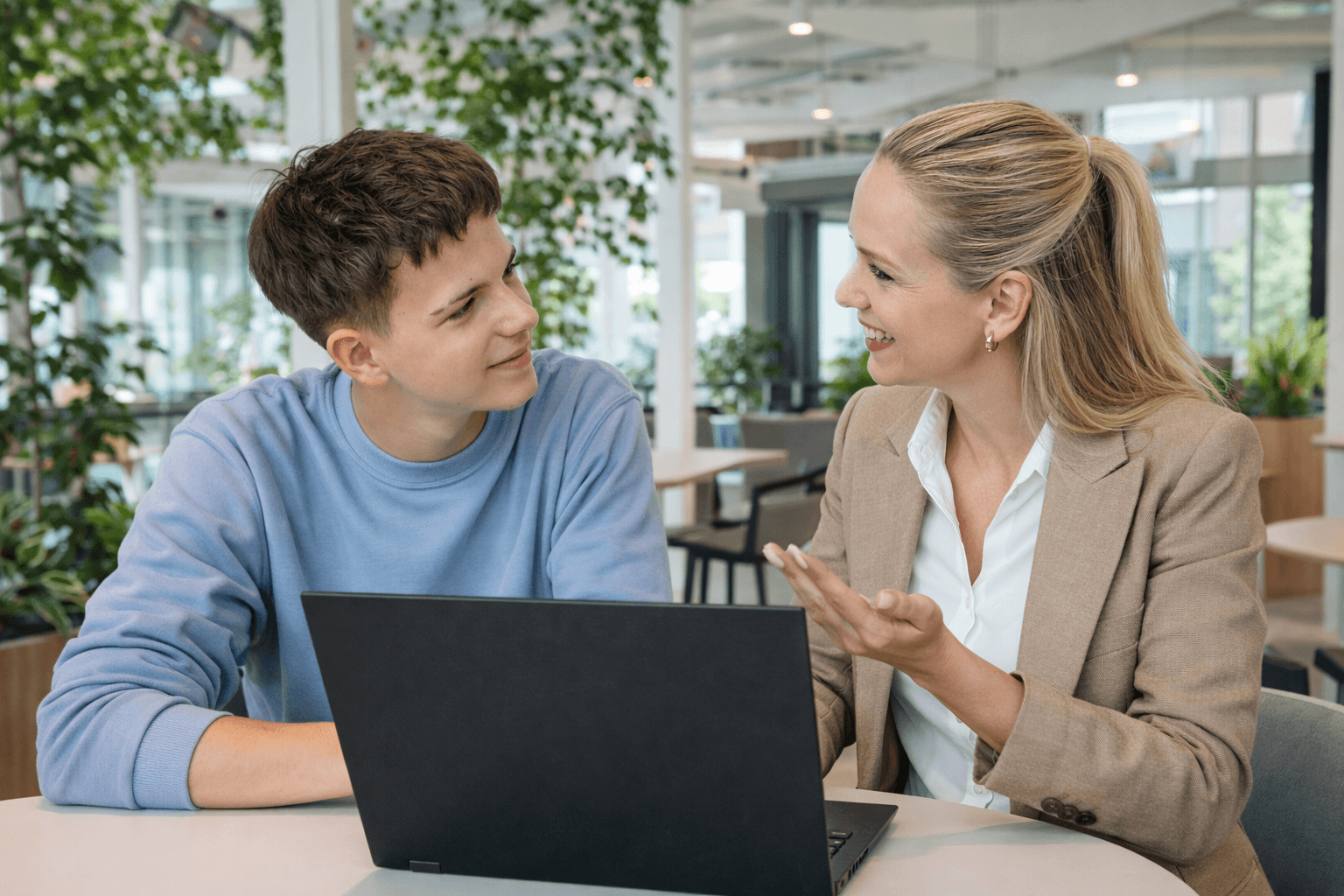 A young person with curly hair smiles at the camera while sitting in front of a laptop against a dark background.