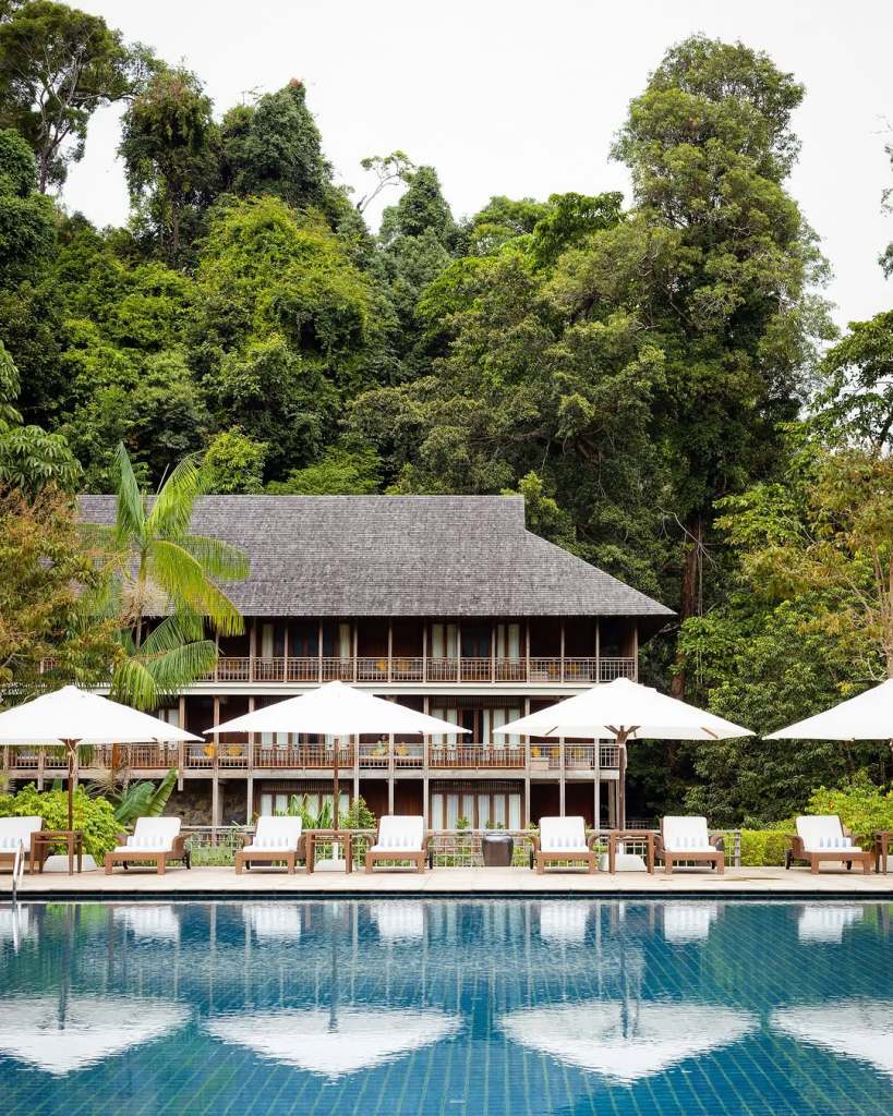 A tranquil view of a resort with wooden architecture surrounded by lush greenery, featuring a swimming pool and sun loungers under white umbrellas.