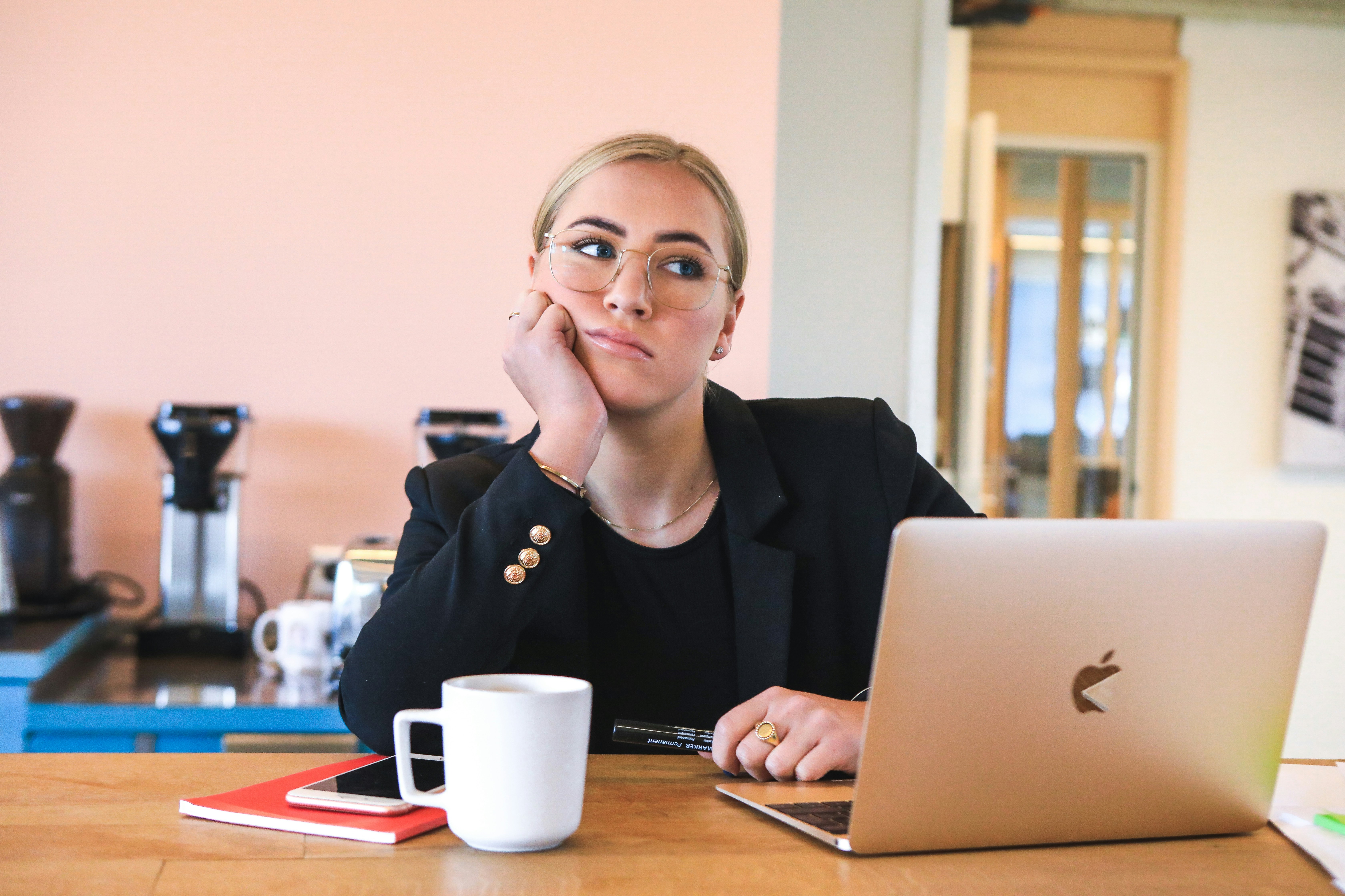 A woman in a blazer sits at a table with an open laptop in front of her. She is resting her head on her fist and looks fed up.