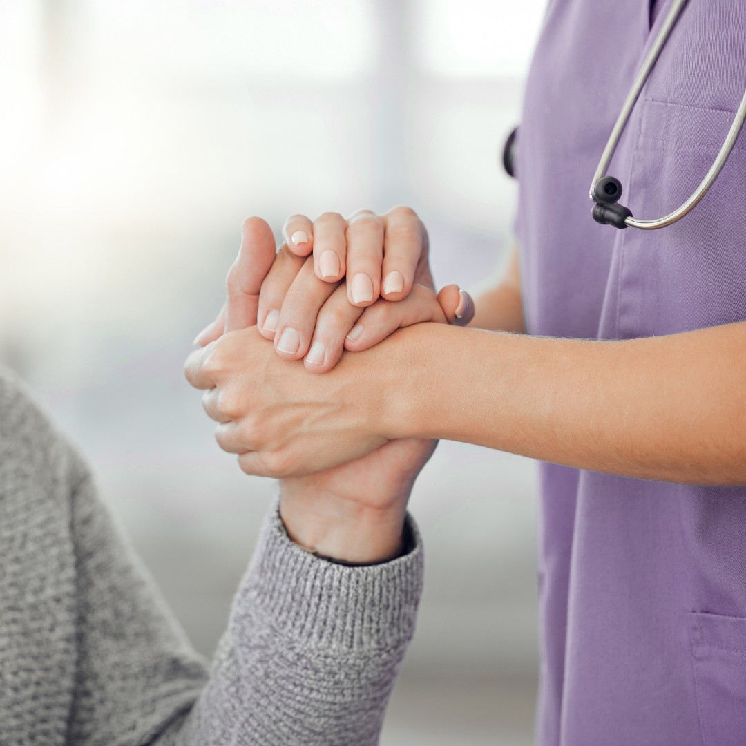 A healthcare professional in a purple uniform gently holds a patient's hand, conveying support and compassion in a clinical setting.