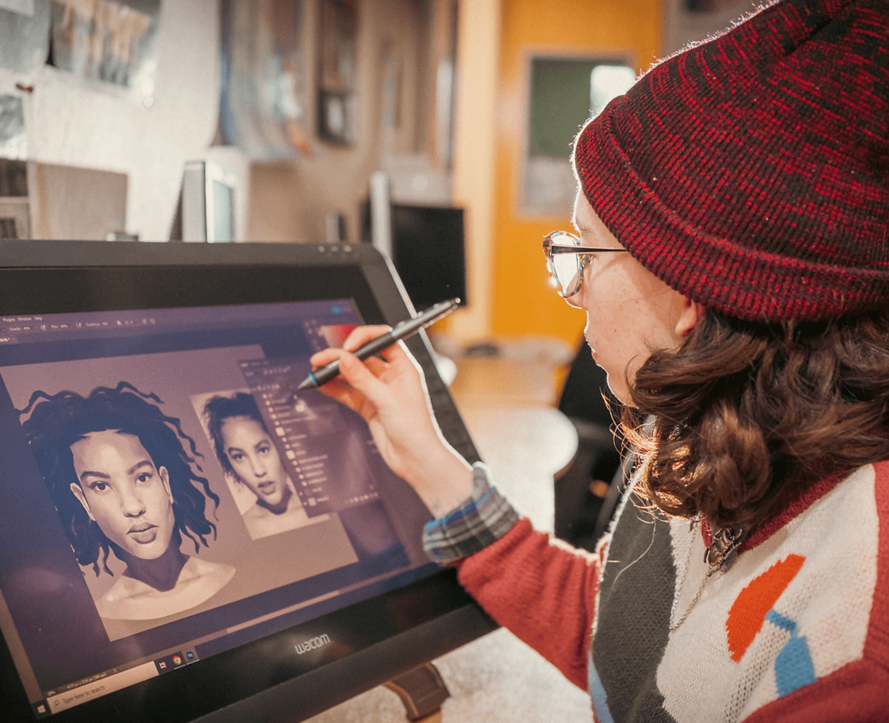 A person wearing a red beanie and glasses is using a digital pen on a Wacom tablet to create a detailed portrait of a woman on the screen, surrounded by a cozy, artistic workspace.