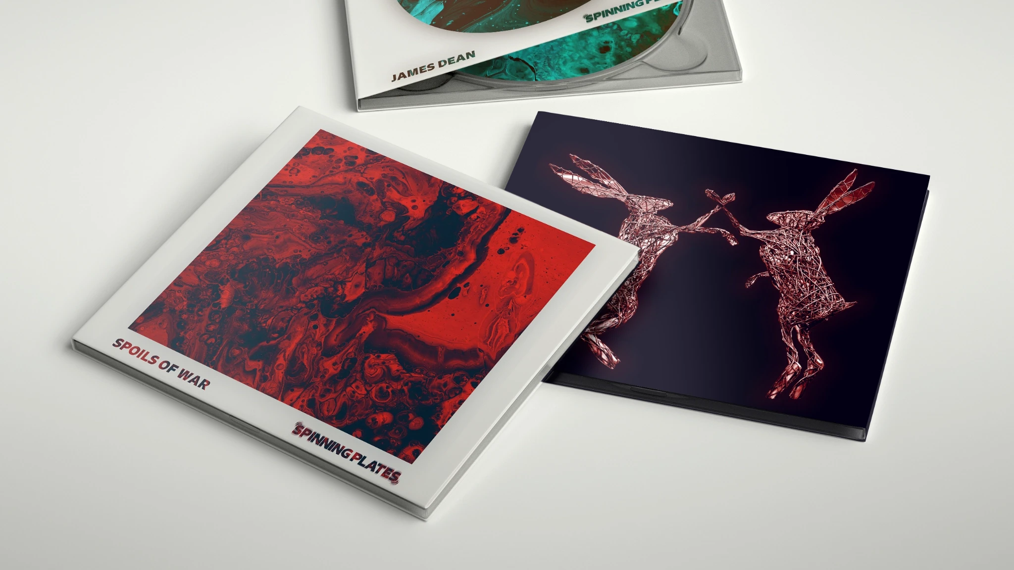 Three albums lying on a surface - one featuring an aqua paint-poured circle (James Dean), one featuring a red paint-poured square (Spoils of War), and one featuring two wire rabbits fighting.