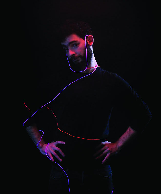 A man with dark hair and a beard stands against a black background. He wears a black shirt and dark pants. Pink and purple neon lights trace his neck and shoulders, extending upward and outward. His hands rest on his hips, and he looks toward the left side of the frame.