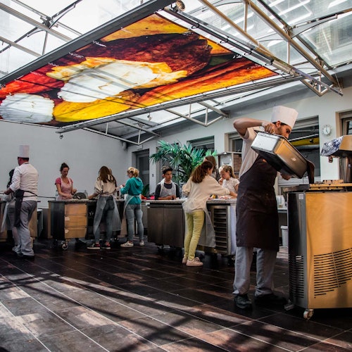 Emberek főznek egy modern konyhában, séfek segítenek nekik, nagy színes, étel témájú mennyezeti falfestmény alatt.