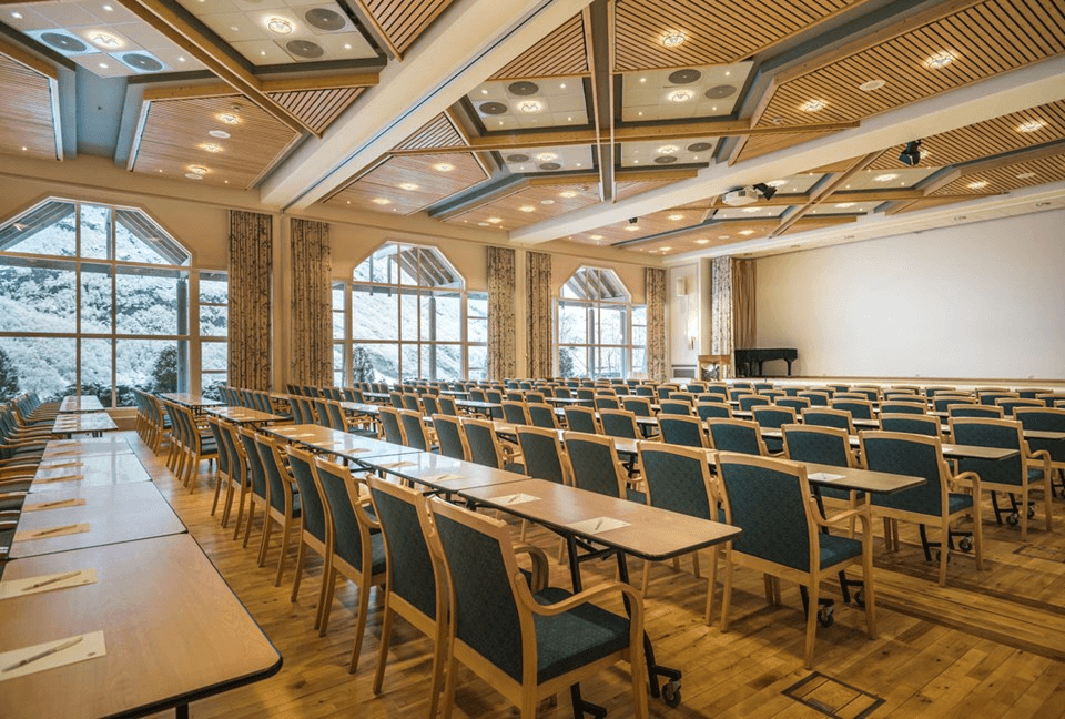 Spacious conference room with rows of wooden tables and blue cushioned chairs, large windows allowing natural light to illuminate the space, and ceiling lights for additional brightness, creating an ideal setting for professional meetings or seminars.