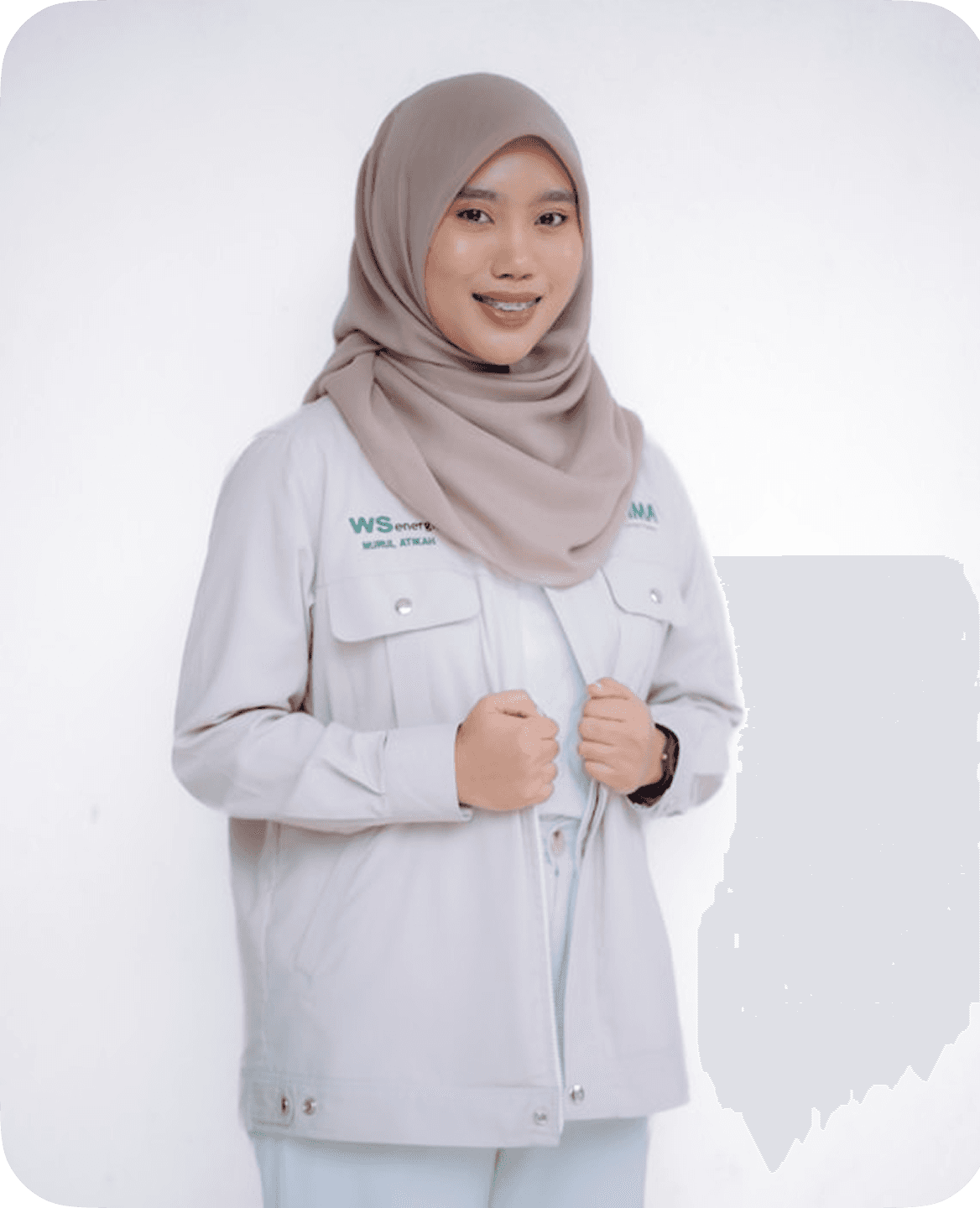 A smiling woman wearing a neutral-toned hijab and light gray jacket with text on it, standing against a plain white background.