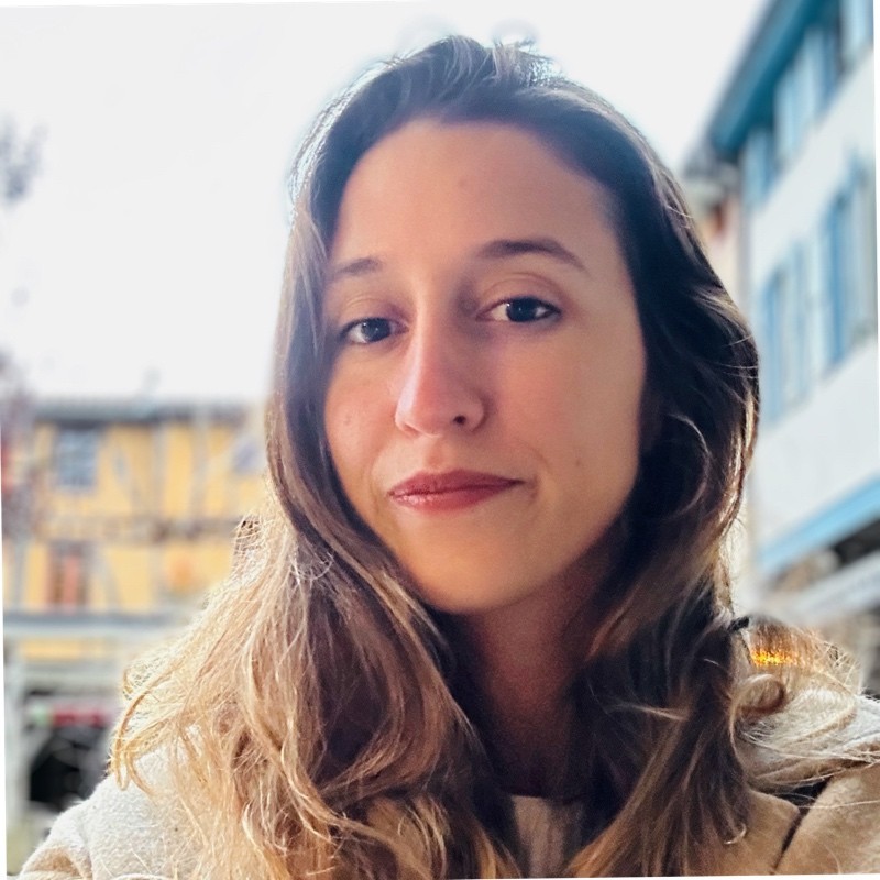 Person in denim shirt over striped tee standing outdoors with blurred buildings in the background
