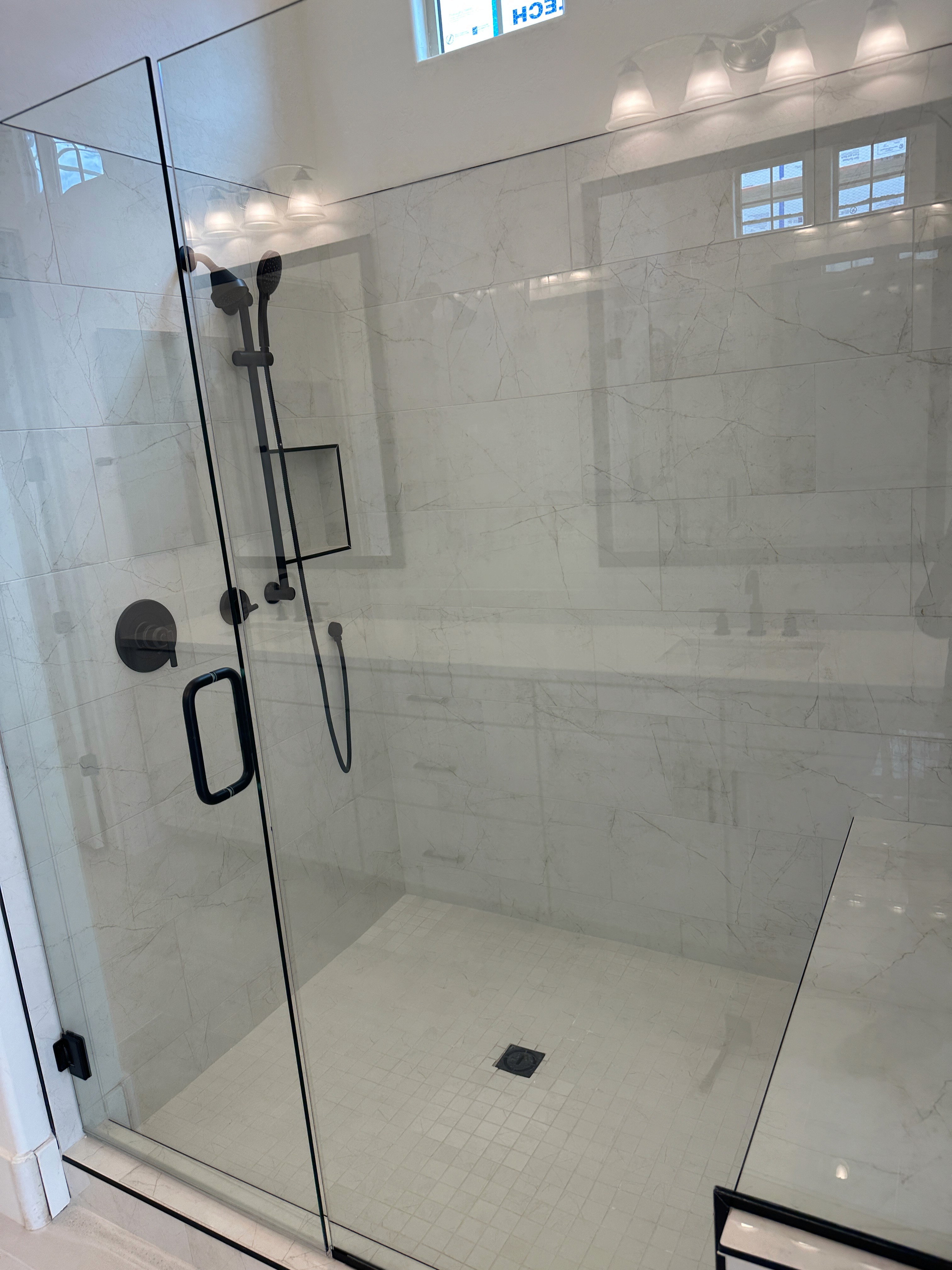 Spacious walk-in shower with clear glass enclosure and black fixtures, featuring white marble-look wall tiles and a white large format tile shower floor, part of a contemporary bathroom remodel.
