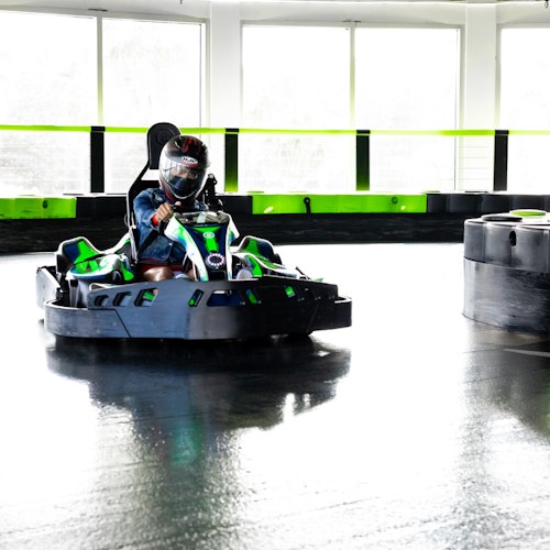 Лице, управляващо зелено и черно картинг на вътрешна писта с отразяващ под и големи прозорци на заден план.