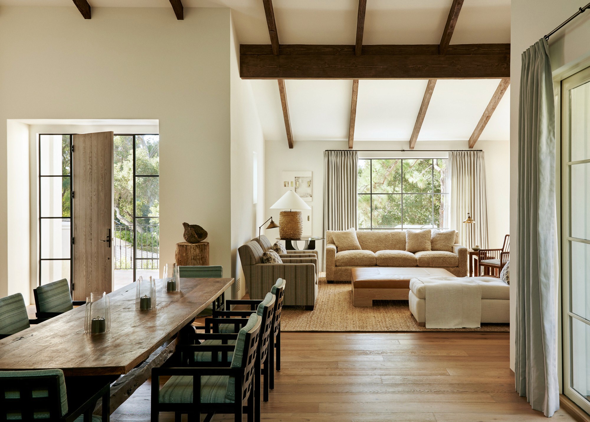 A view through the dining area into the living room beyond. A rustic trestle table is surrounded by dark-framed chairs with green upholstery. The living room features layered seating on a jute rug, with exposed beams overhead and steel-and-wood French doors open to the garden.
