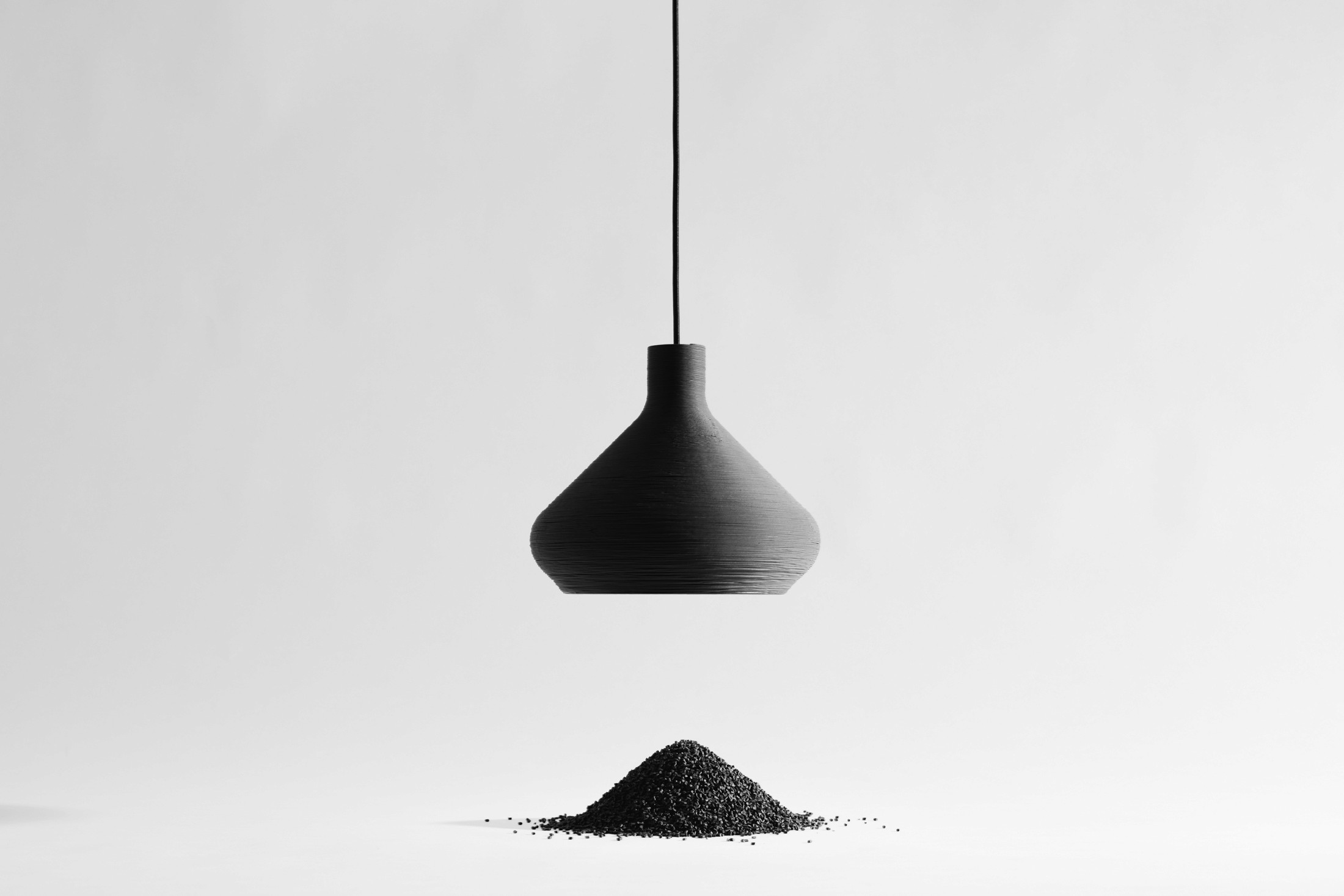 A black pendant light fixture against a white background, with a pile of black pellets beneath it