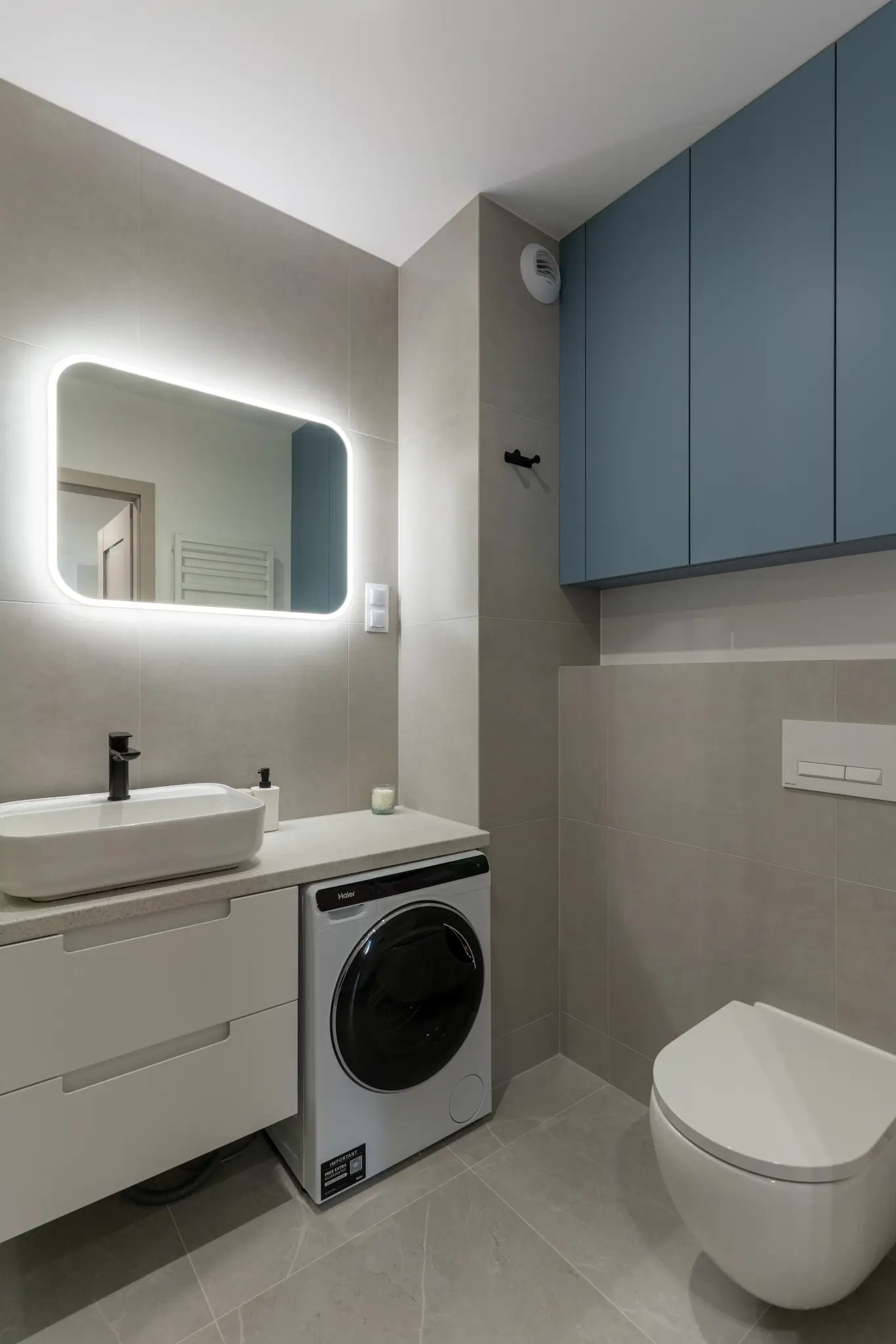 Nowoczesna mała łazienka z podświetlanym lustrem, białą szafką pod umywalkę i zabudową nad pralką.