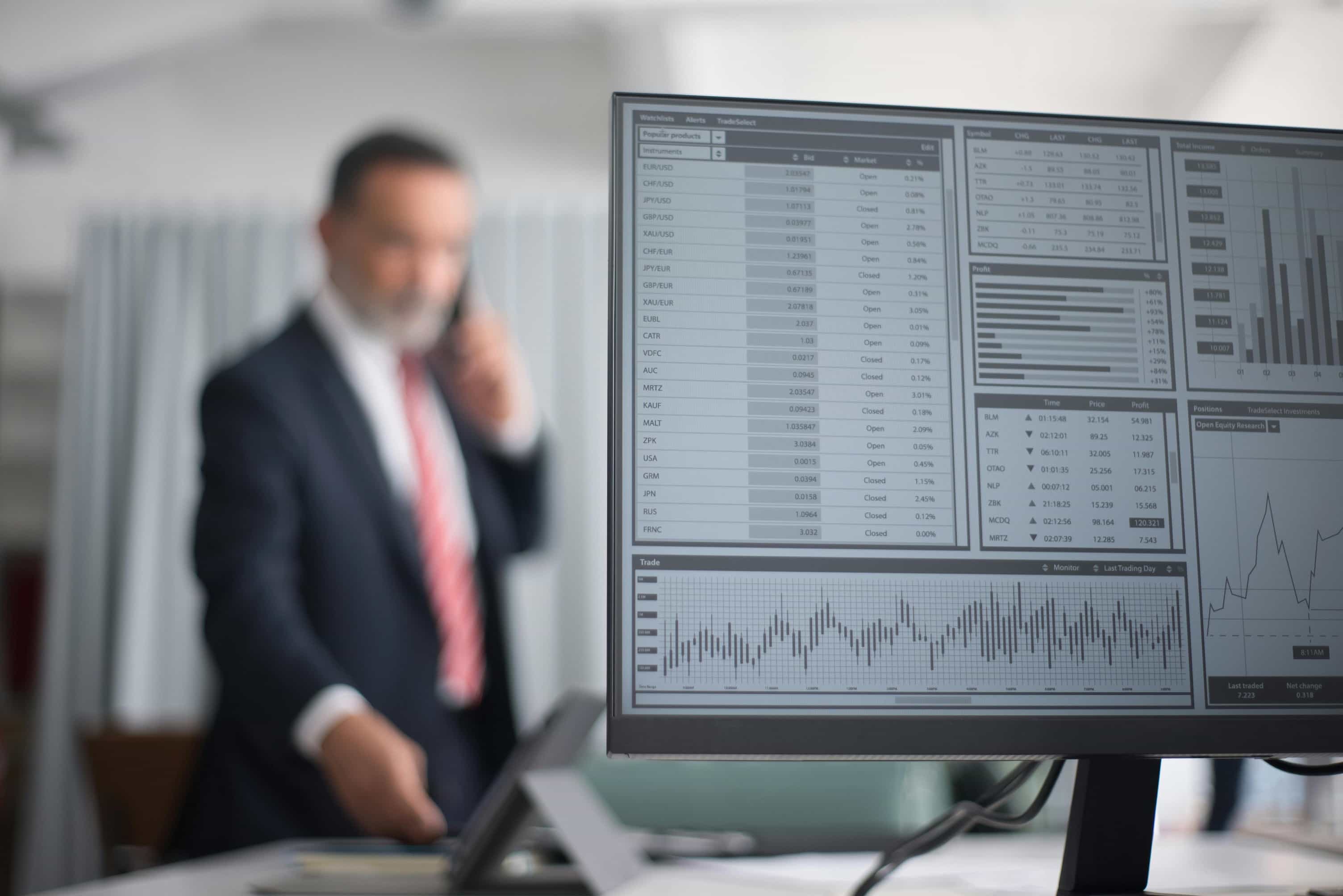 A close-up of a computer monitor displaying financial graphs, charts, and data tables. In the blurry background, a man in a dark suit and red tie is talking on a phone.
