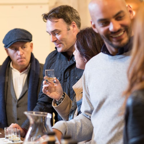 People standing at a counter, holding glasses of whiskey, engaged in conversation. One person wears a cap; another smiles.