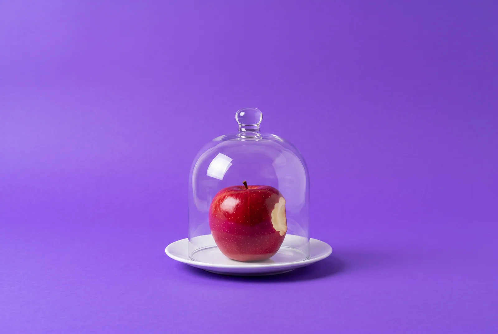 Red apple with one bite taken out, resting under a clear glass dome on a lilac backdrop, illustrating the best Christian app to quit porn