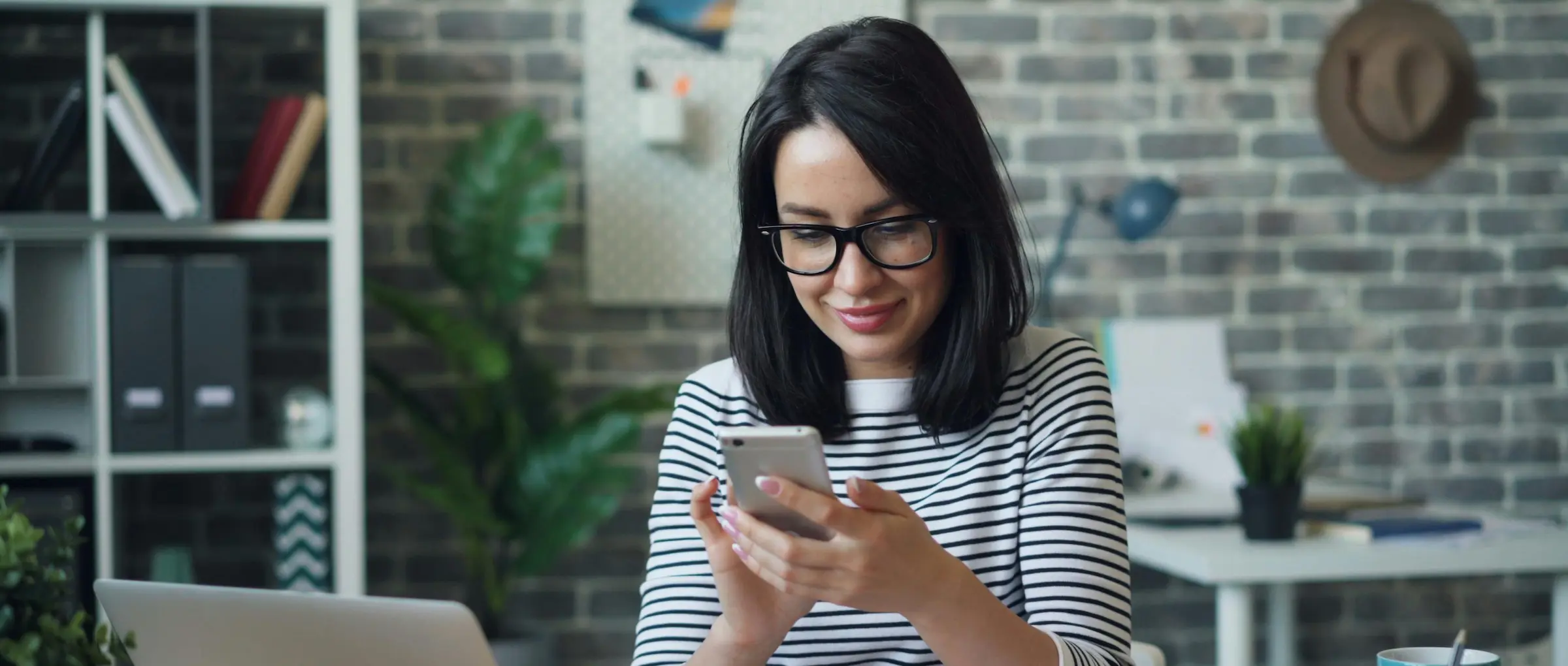 A woman sitting at a desk smiling while using her phone, with a laptop and home office setting in the background.