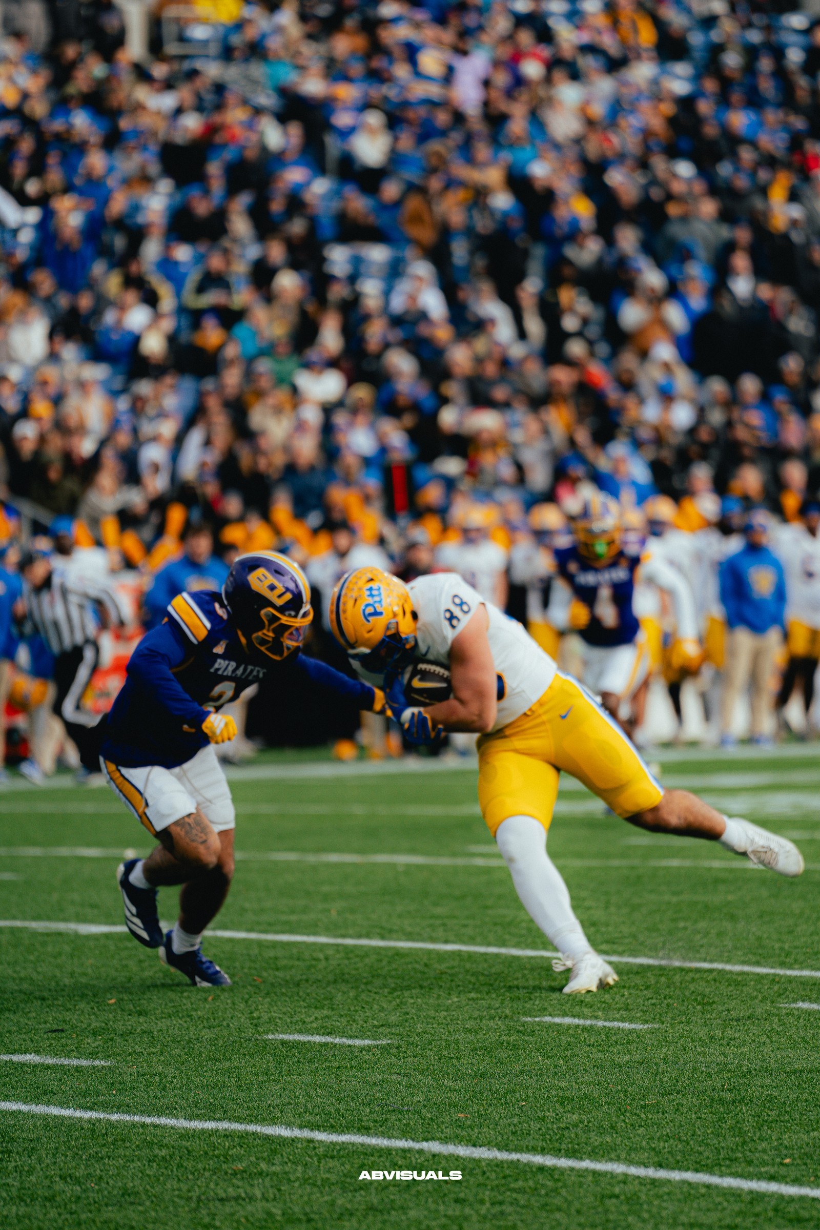 A football ball carrier in a white jersey and yellow pants leans forward into contact with a blue-and-yellow-uniformed defender.